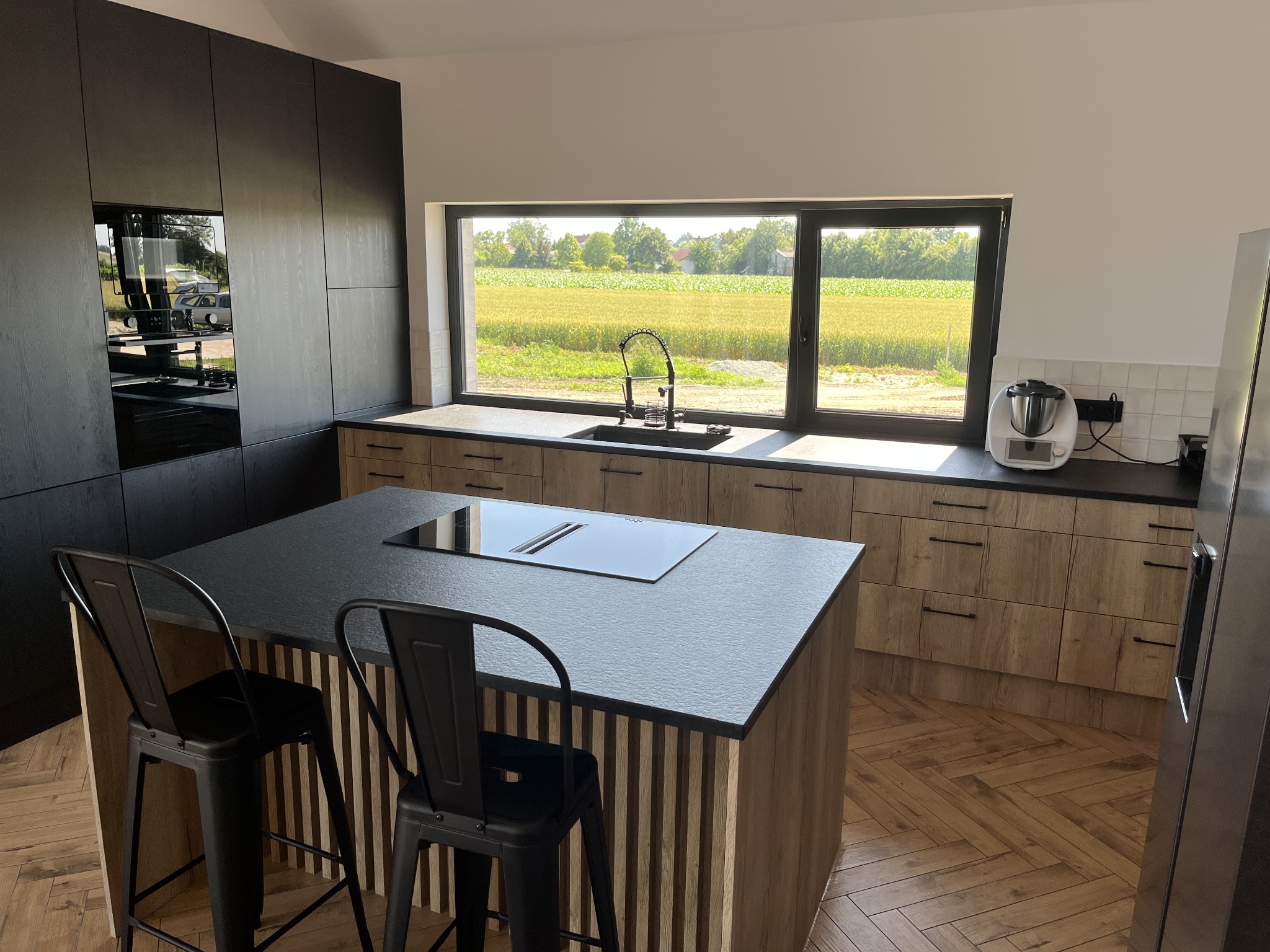 Nowoczesna wyspa kuchenna z blatem kamiennym w Ciepłowodach. Czarny blat, drewniane wykończenie, metalowe krzesła barowe. Widok na pole z okna.