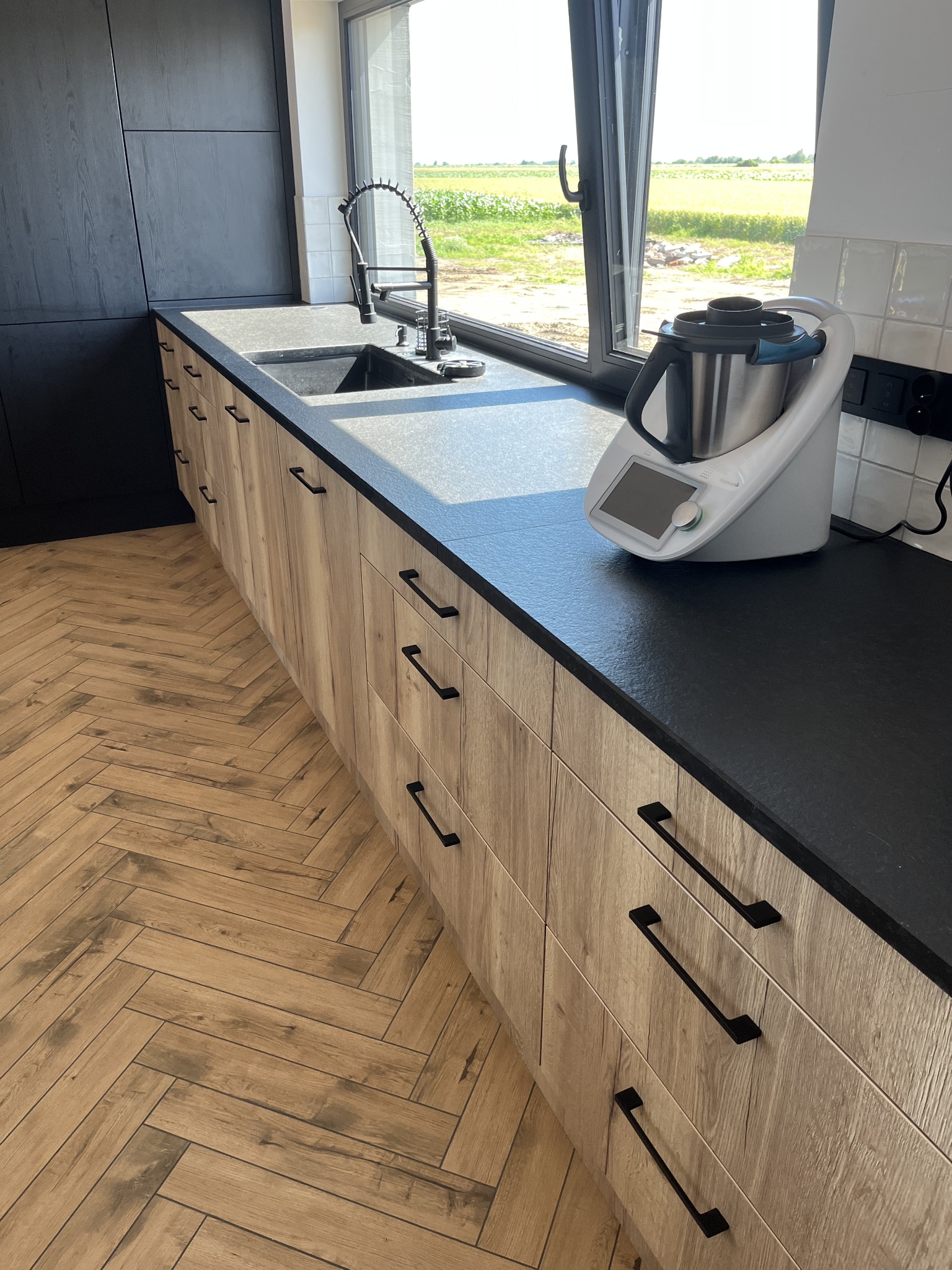 Czarny blat kamienny w nowoczesnej kuchni z drewnianymi szafkami i widokiem na pole za oknem. Bateria kuchenna i robot kuchenny na blacie.