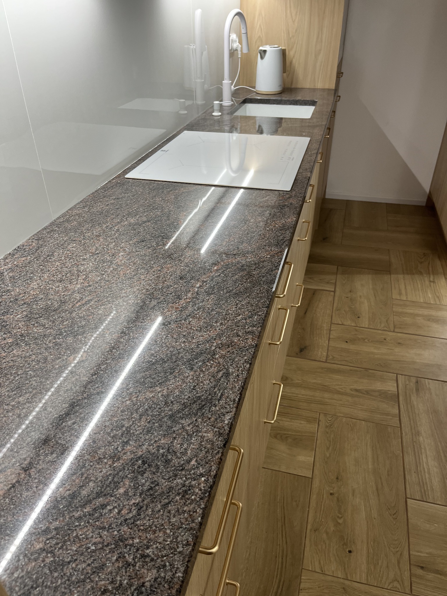 Błyszczący, brązowo-szary blat kamienny w kuchni z płytą indukcyjną i zlewozmywakiem. Złote uchwyty szafek i drewniana podłoga w jodełkę.