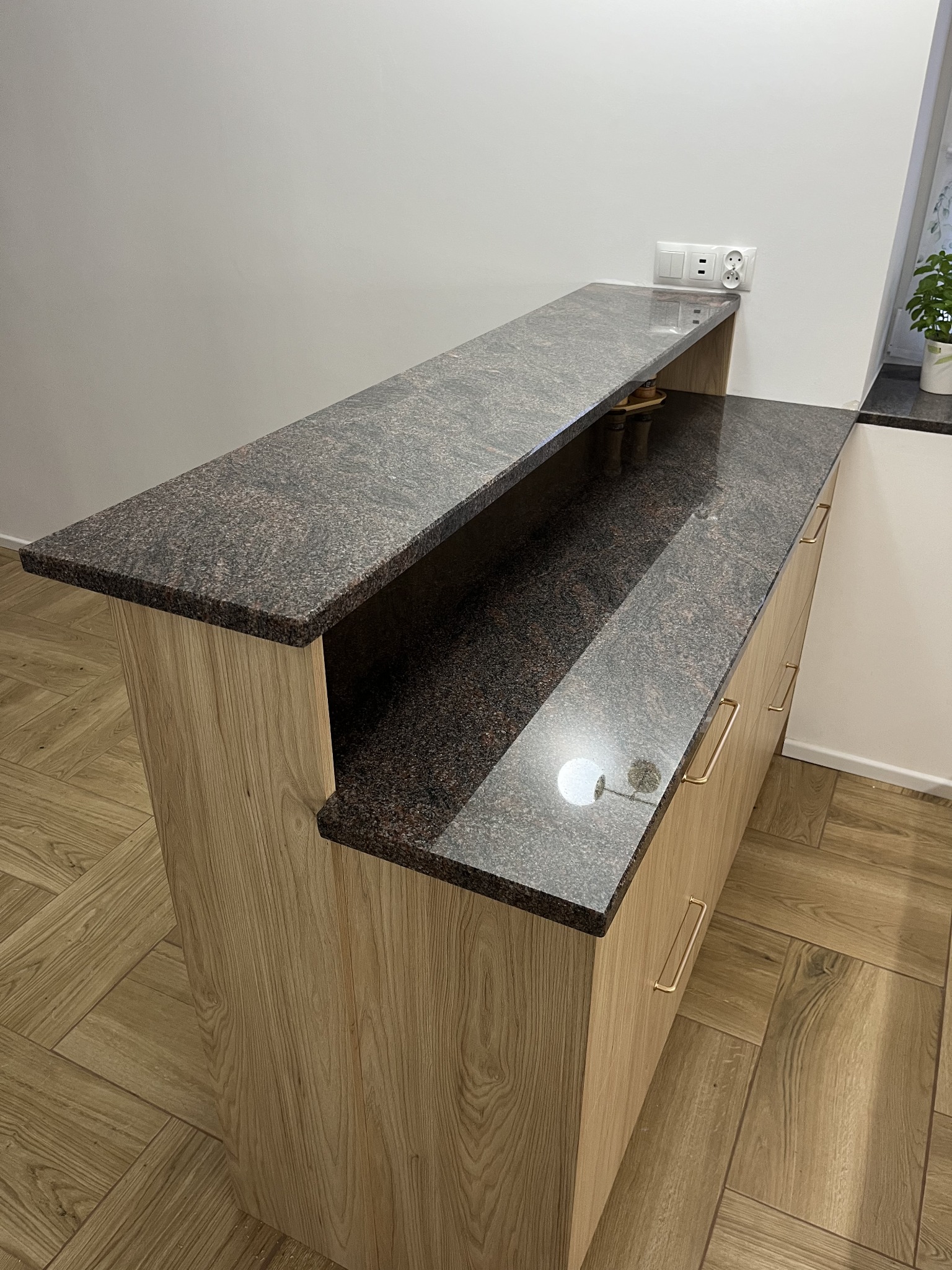Granitowy blat kuchenny z drewnopodobną obudową, widok z perspektywy. Ciemny, nakrapiany kamień, złote uchwyty szuflad, gniazdko elektryczne na ścianie.