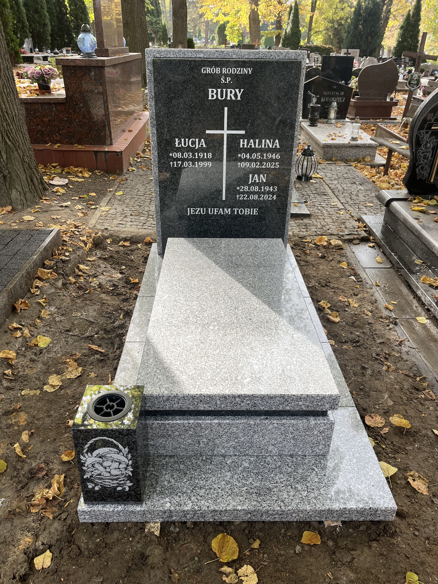 Granitowy nagrobek rodzinny z inskrypcjami i wazonem z motywem grzybów, otoczony jesiennymi liśćmi na cmentarzu.