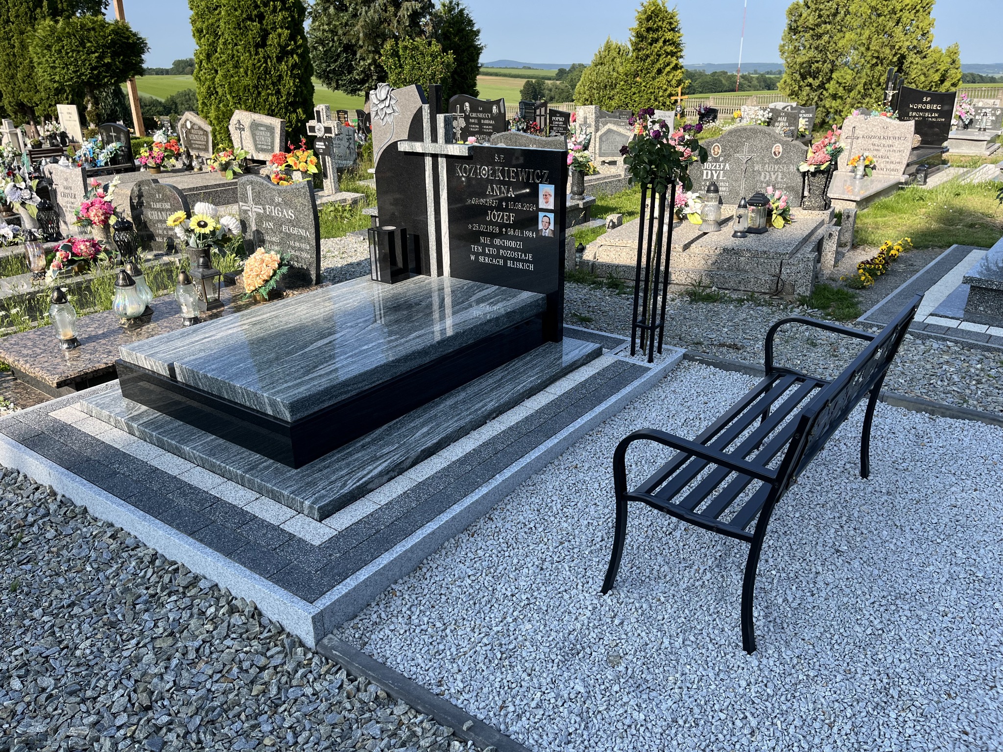 Granitowy nagrobek z ławką na cmentarzu, otoczony białym żwirem. Widok na inne nagrobki i zieleń w tle, słoneczny dzień. Nagrobek z czarną tablicą i zdjęciami.