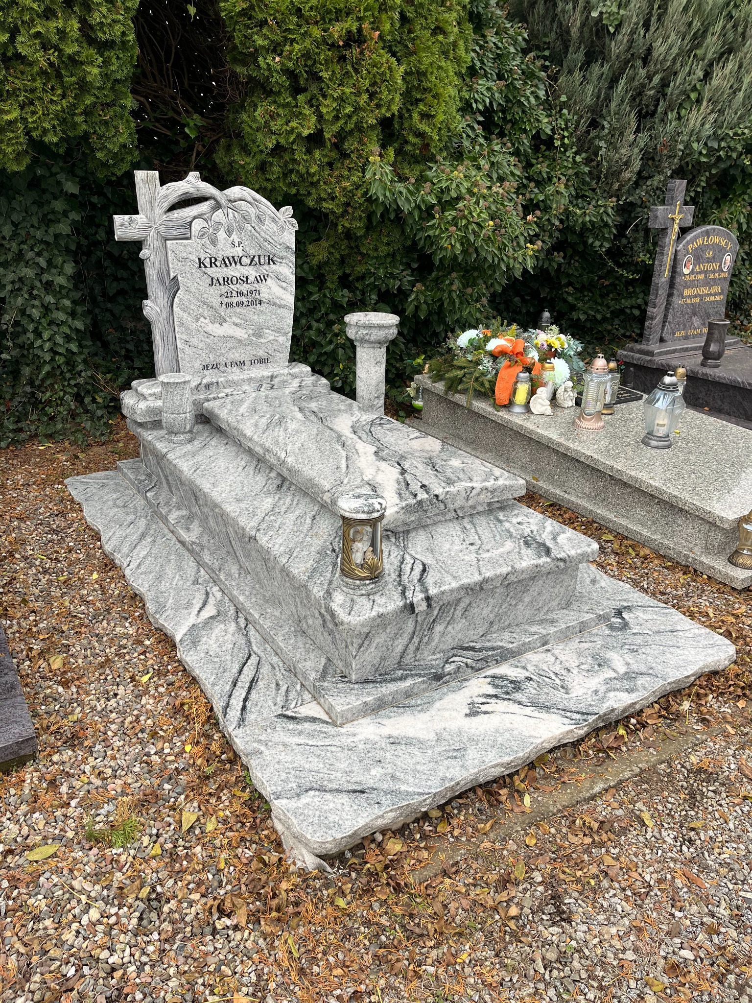 Granitowy nagrobek z krzyżem i inskrypcją 'Krawczuk Jarosław' na cmentarzu, otoczony zielenią. Widoczne znicze i kamienny postument.
