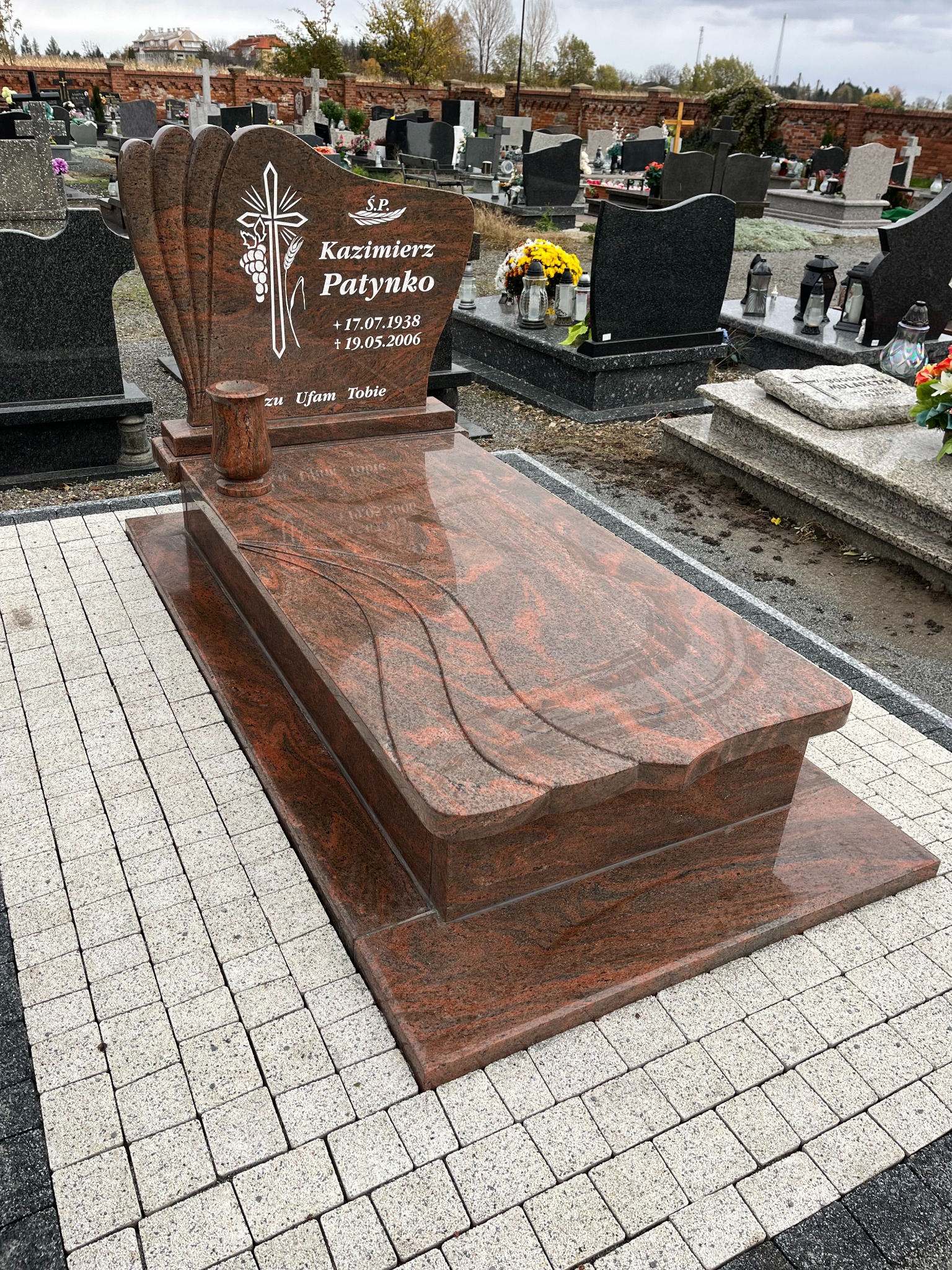 Granitowy nagrobek z inskrypcją 'Kazimierz Patynko' na cmentarzu, otoczony kostką brukową. Widoczne inne nagrobki w tle. Ujęcie z perspektywy.