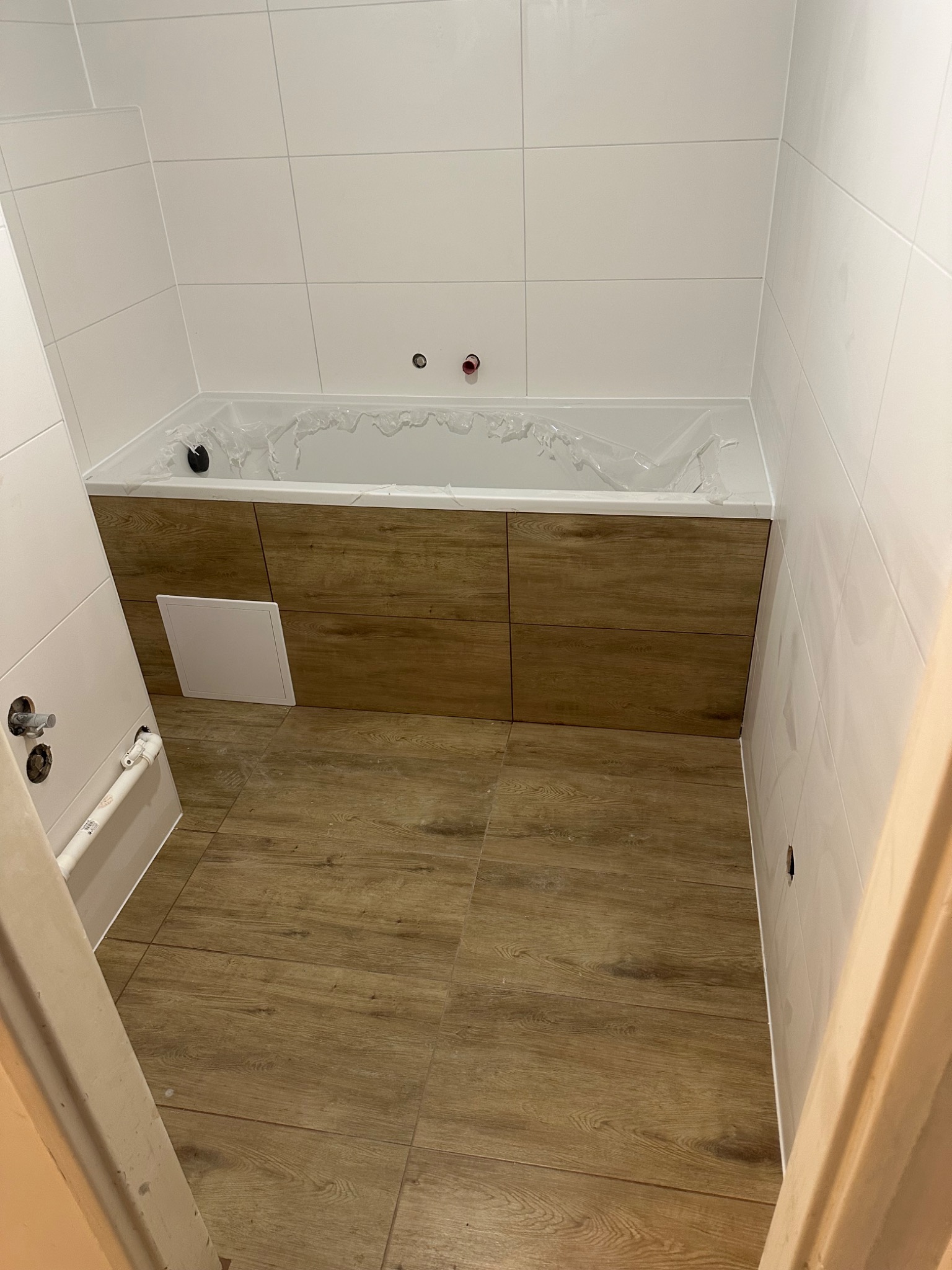 Wykończona łazienka w Poznaniu: białe płytki ścienne, drewnopodobna zabudowa wanny i podłoga z paneli. Widoczne przyłącza wodne i drzwi wejściowe.