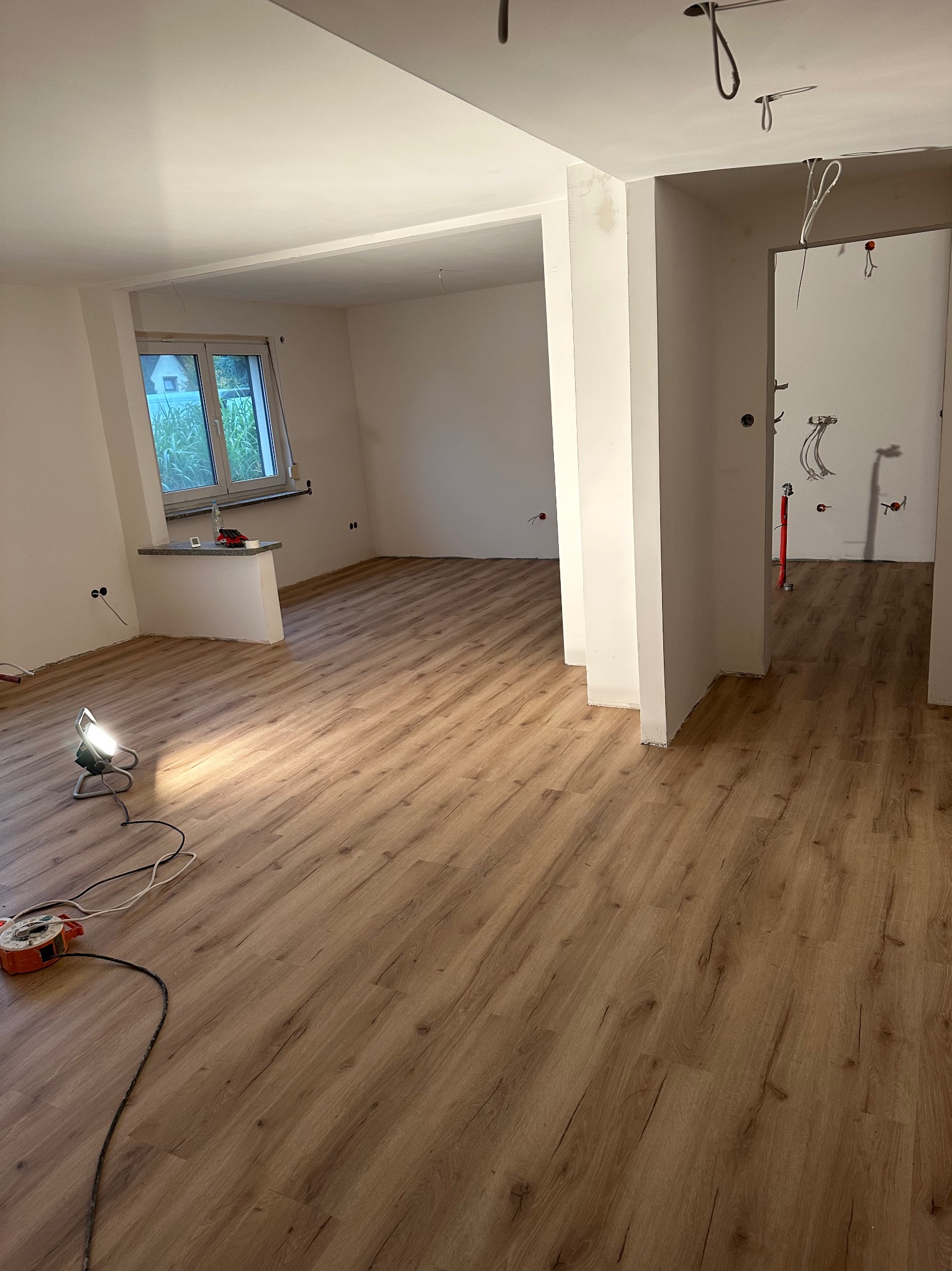 Jasne panele podłogowe w nowym wnętrzu z białymi ścianami i oknem. Widoczne narzędzia pracy i lampa halogenowa. W tle instalacja elektryczna.