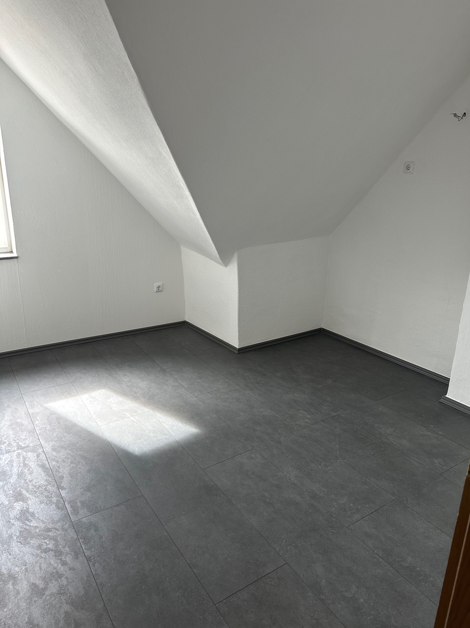 Pokój na poddaszu po remoncie: szare płytki podłogowe, białe ściany z fakturą, skosy. Widoczne gniazdka elektryczne i listwy przypodłogowe. Wpadające światło słoneczne.