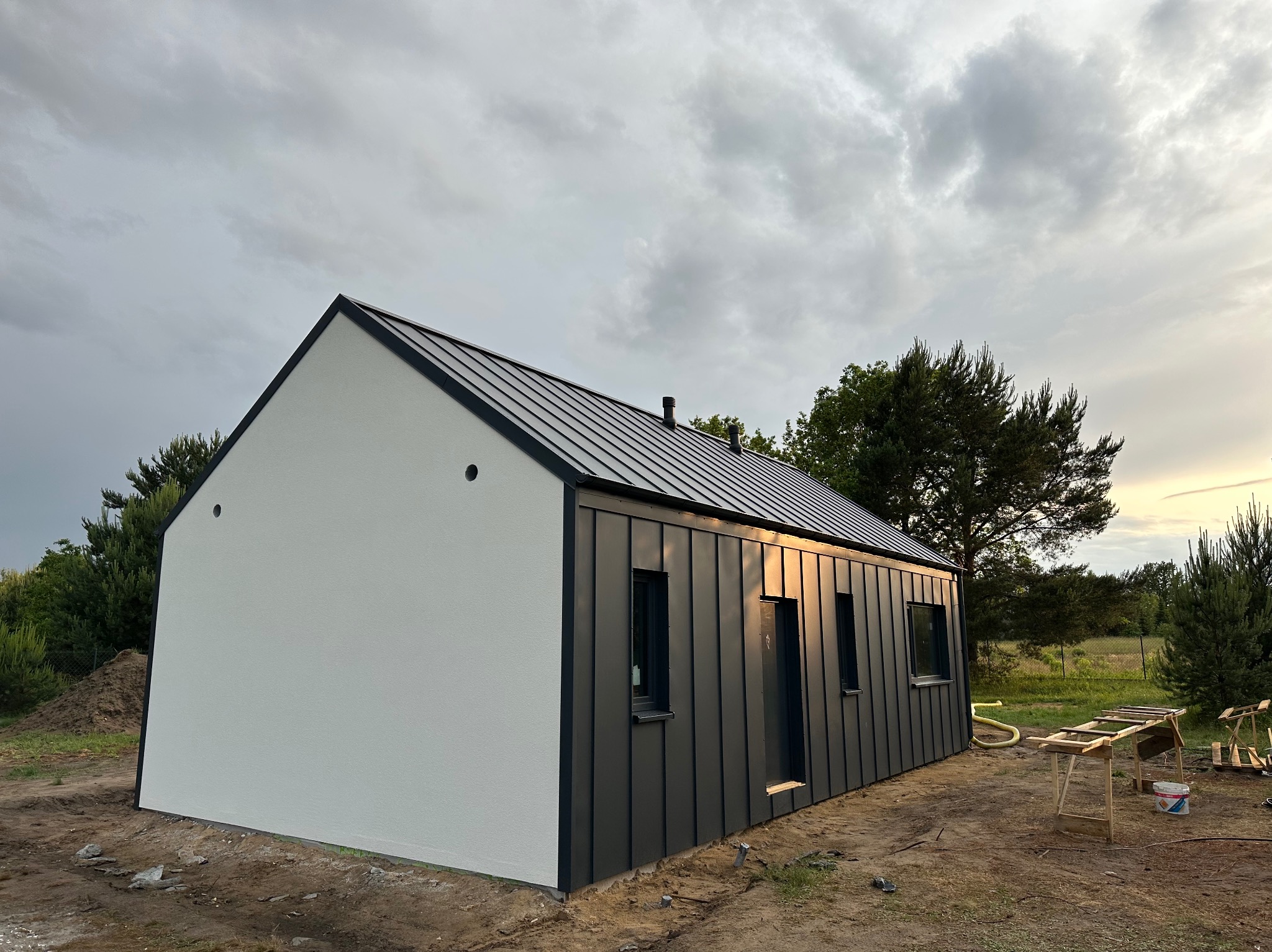 Nowoczesny dom jednorodzinny z białą elewacją i grafitową blachą na rąbek stojący, widok z zewnątrz, w tle zachmurzone niebo i zieleń. Częściowo zagospodarowany teren.