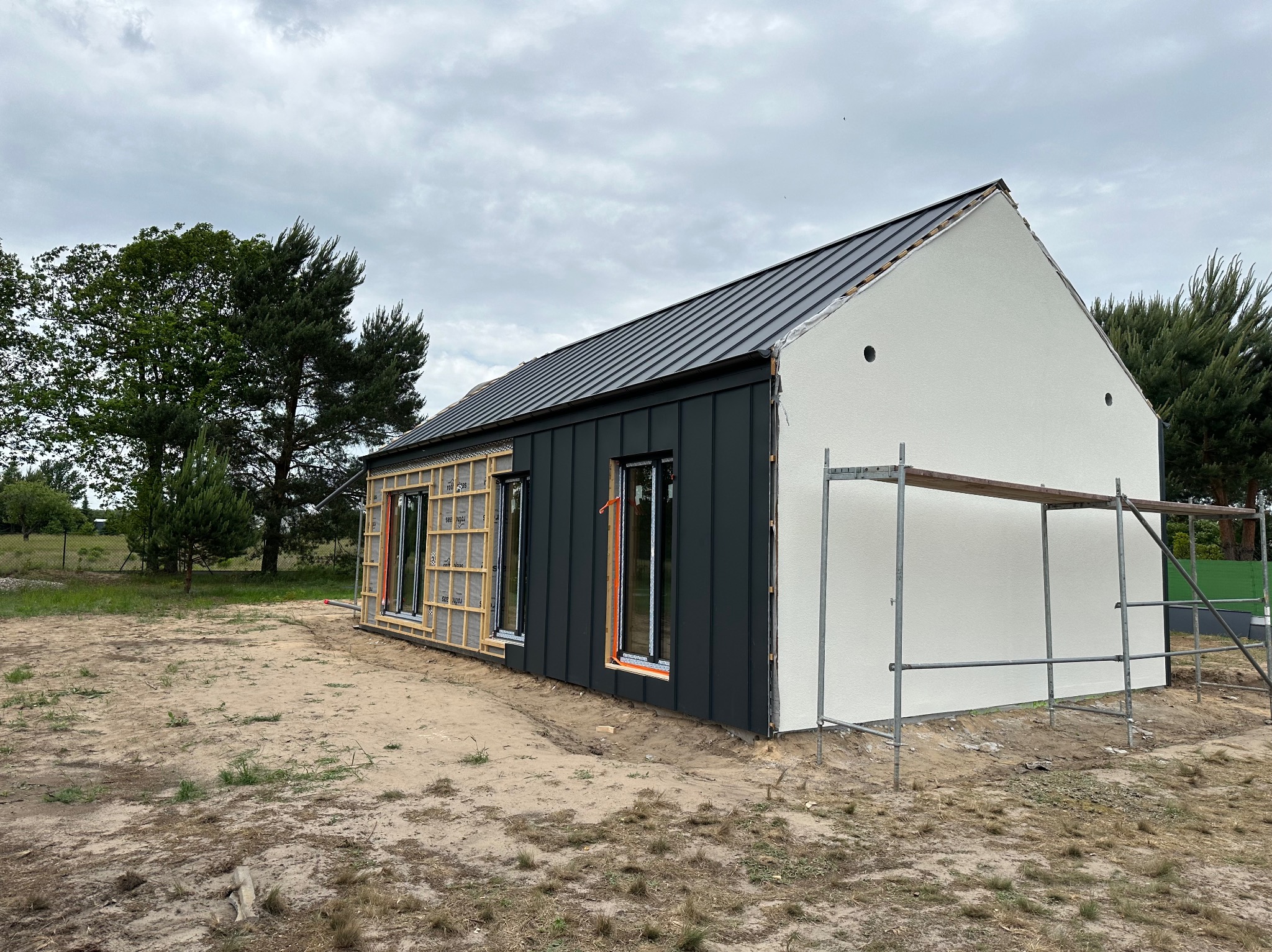 Dom w trakcie budowy: część elewacji otynkowana na biało, część z czarną blachą trapezową, widoczne rusztowanie i okna w surowej ramie.