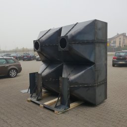 Teknequip - Duży, spawany, metalowy element konstrukcyjny o geometrycznym kształcie, z otworami, umieszczony na drewnianych paletach na zewnątrz, na tle parkingu i budynków.