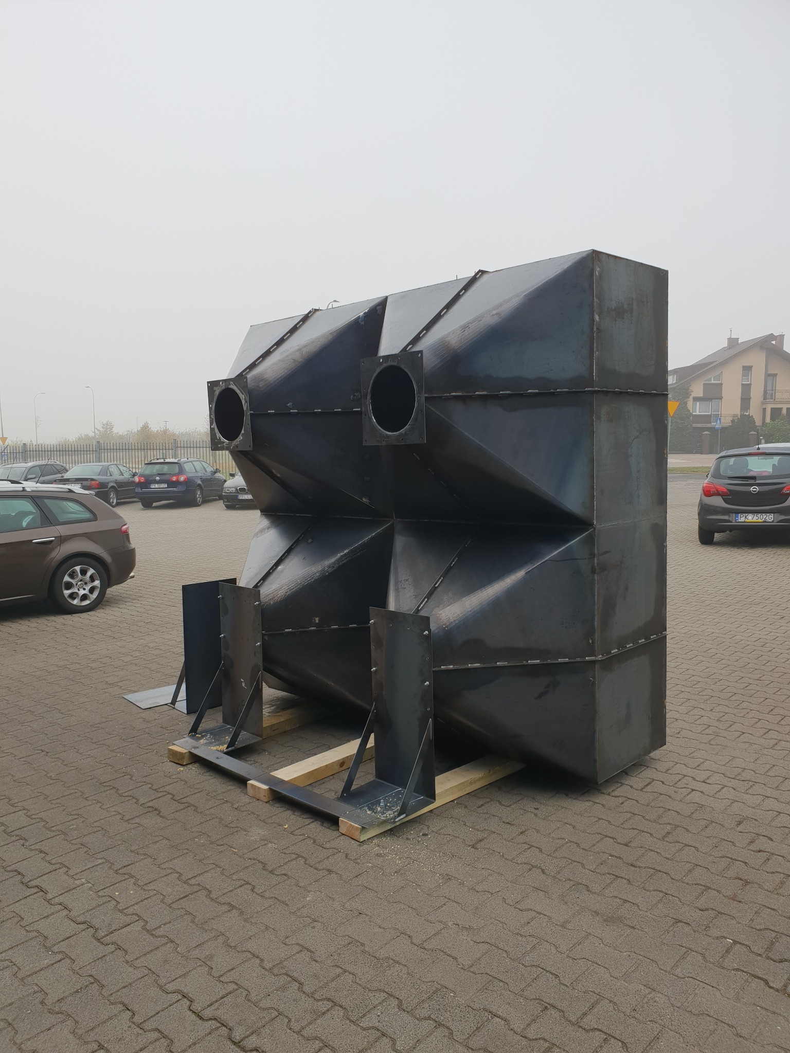 Duży, spawany, metalowy element konstrukcyjny o geometrycznym kształcie, z otworami, umieszczony na drewnianych paletach na zewnątrz, na tle parkingu i budynków.