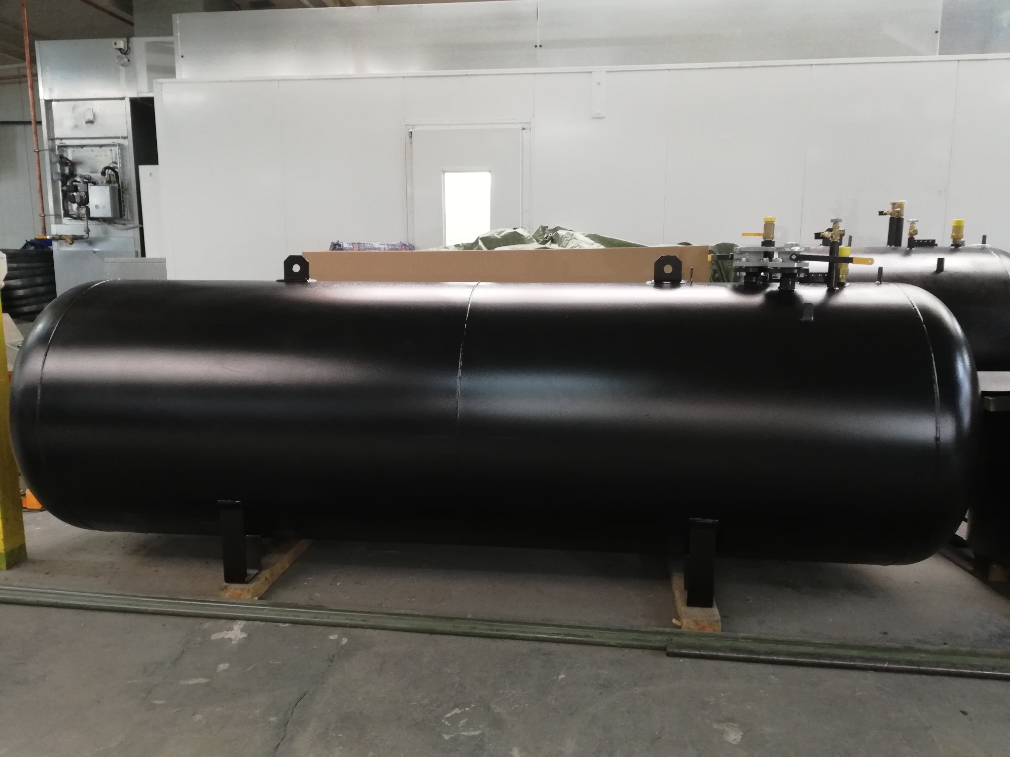 Czarny, poziomy zbiornik ciśnieniowy na drewnianych podporach w hali produkcyjnej. Widoczne zawory i elementy instalacji. Drugi zbiornik w tle.