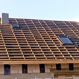 TWYOJ MAJSTER - Konstrukcja dachu z oknami dachowymi w trakcie budowy, widoczne drewniane łaty i membrana. Kominek z szarego pustaka. Ściany budynku z betonu komórkowego.