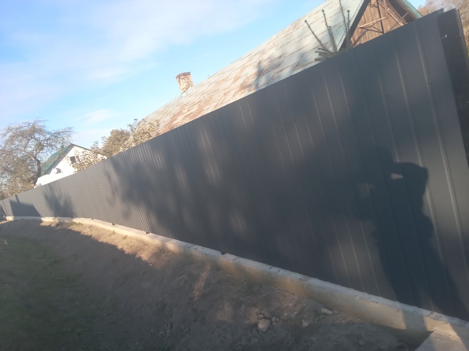 Nowy, grafitowy płot z blachy falistej, oddzielający posesję od drogi. W tle dom z kominem i drzewa. Cień osoby fotografującej widoczny na płocie.