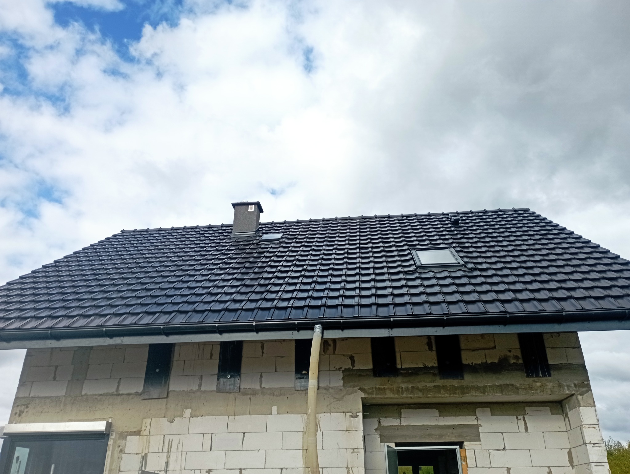 Dom w trakcie budowy z czarną dachówką i widocznym kominem na tle pochmurnego nieba. Widoczne białe bloczki konstrukcyjne i rynna.