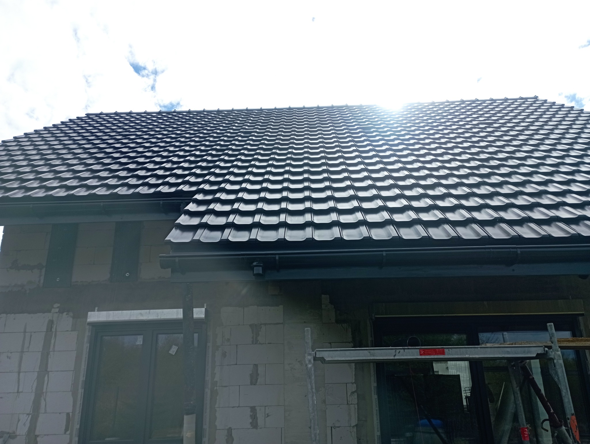 Nowy, błyszczący, czarny dach z blachodachówki na domu w trakcie budowy. Widoczne rusztowania i surowy mur z bloczków.