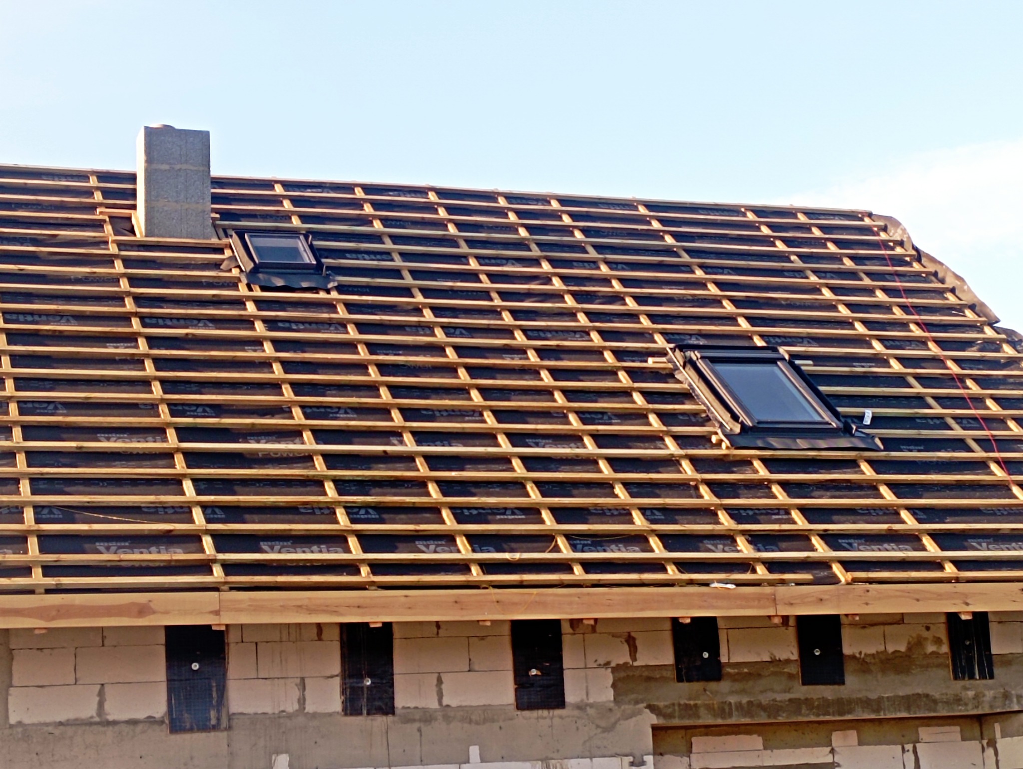 Konstrukcja dachu z oknami dachowymi w trakcie budowy, widoczne drewniane łaty i membrana. Kominek z szarego pustaka. Ściany budynku z betonu komórkowego.