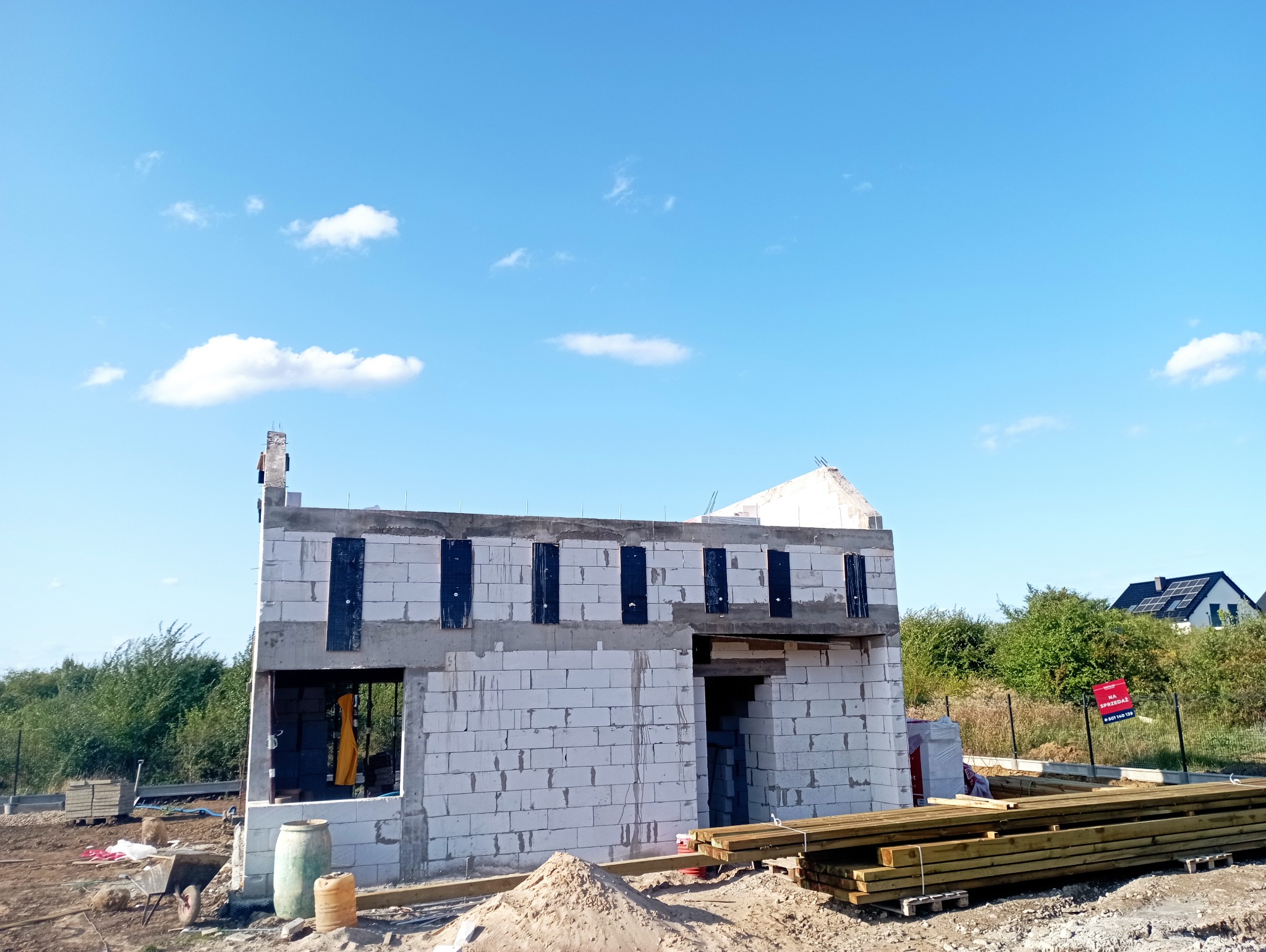 Surowy stan domu w budowie z białych bloczków, widoczne otwory okienne i drzwiowe, czarne elementy izolacyjne, błękitne niebo w tle. Etap wznoszenia ścian.