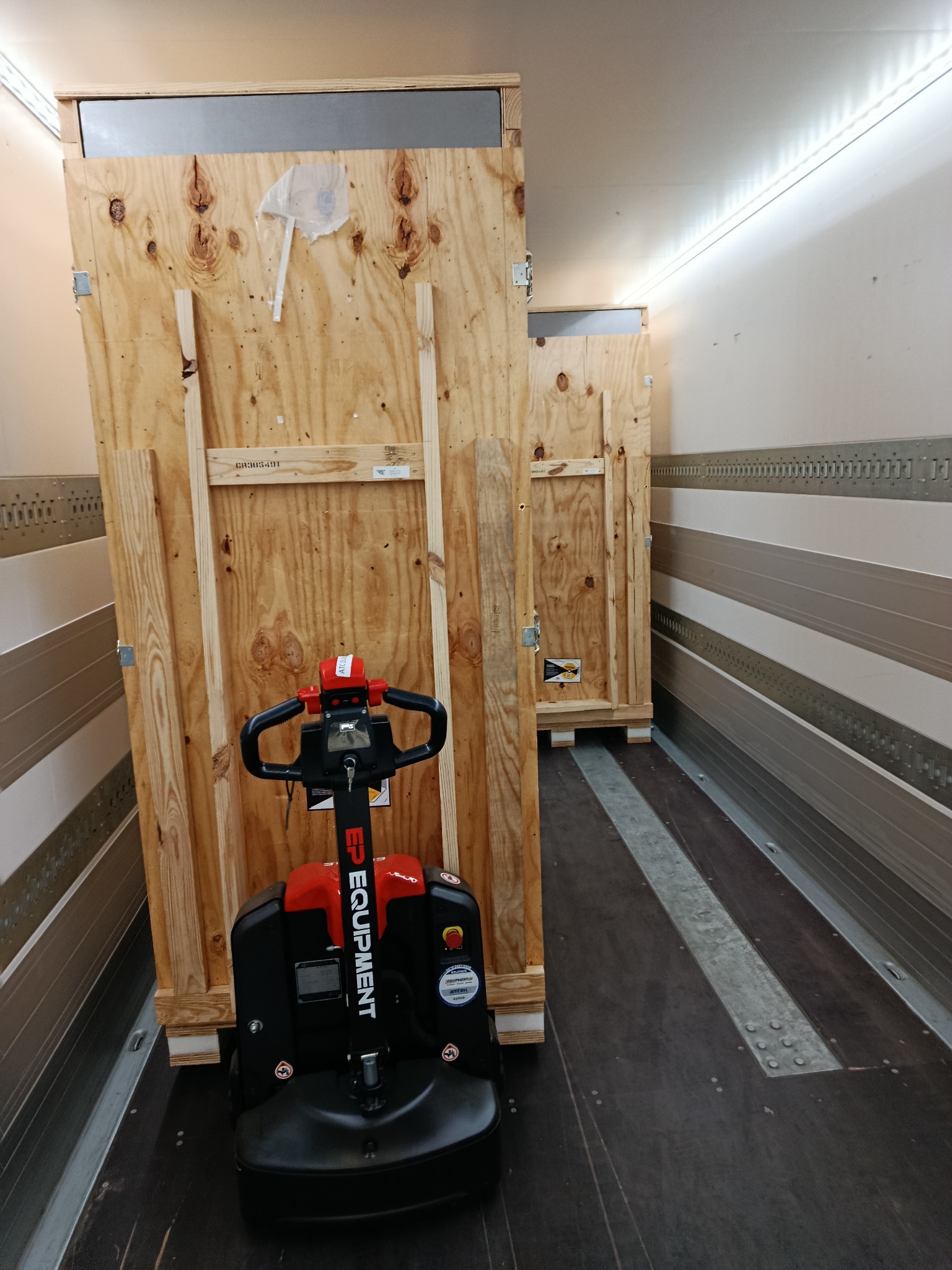 Wnętrze naczepy z paleciakiem elektrycznym i drewnianymi skrzyniami zabezpieczonymi do transportu. Widoczne szyny mocujące ładunek.