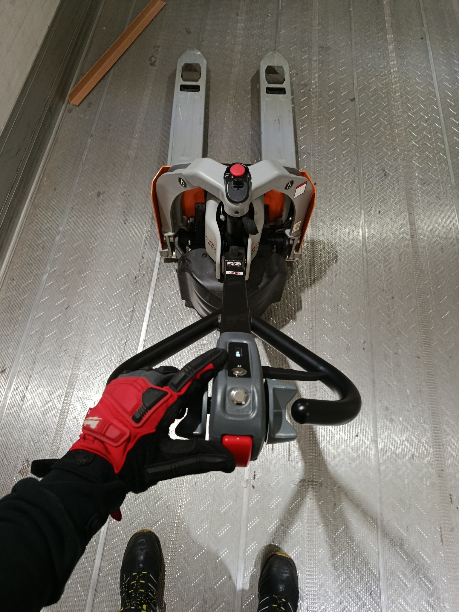 Elektryczny wózek paletowy wewnątrz metalowej naczepy, obsługiwany przez osobę w rękawicach roboczych. Widoczne buty operatora. Podłoga naczepy z blachy ryflowanej.