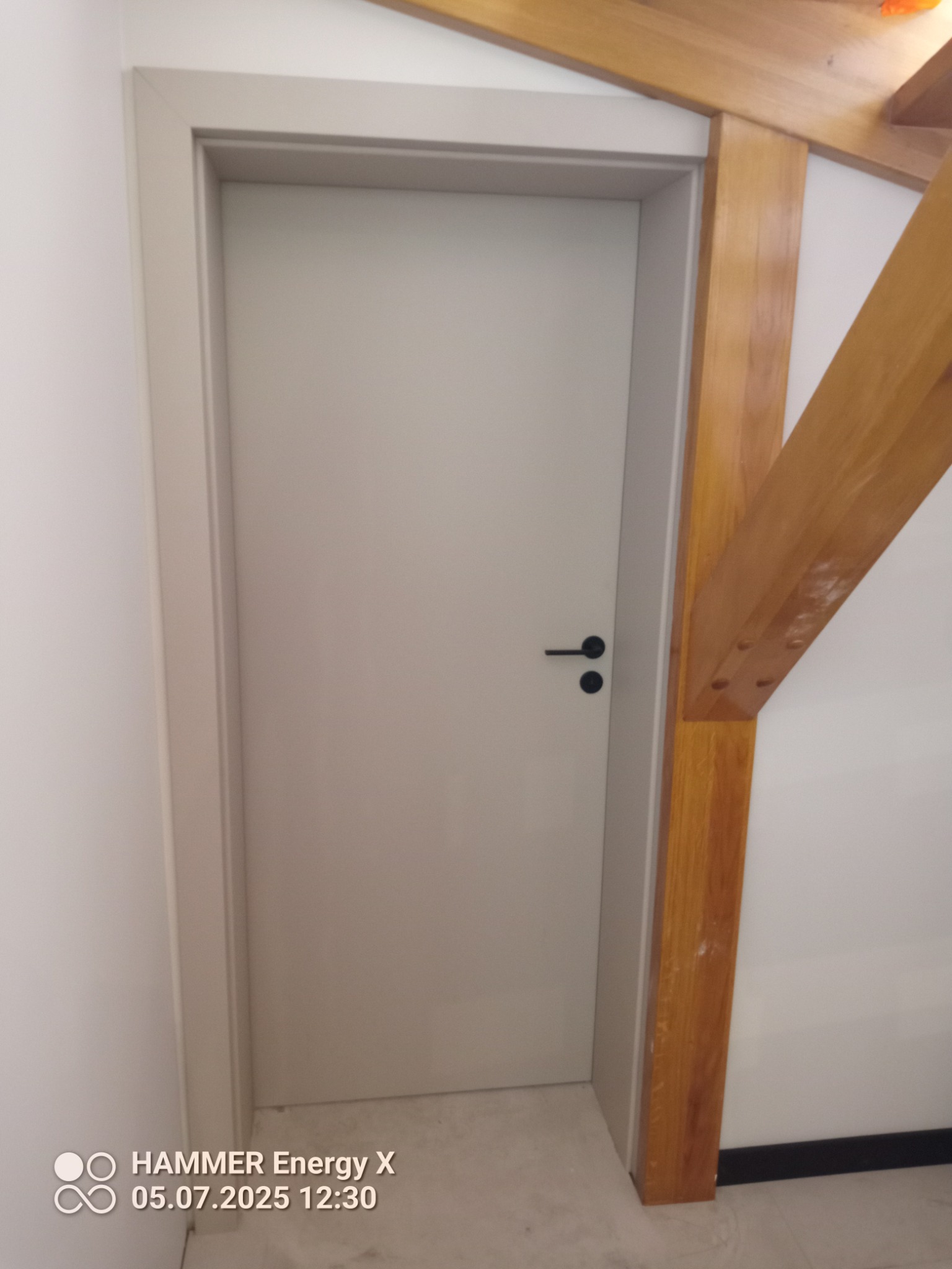 Zamknięte, szare drzwi wewnętrzne z czarną klamką, osadzone w białej ścianie z drewnianą konstrukcją stropu po prawej stronie. Minimalistyczny design.