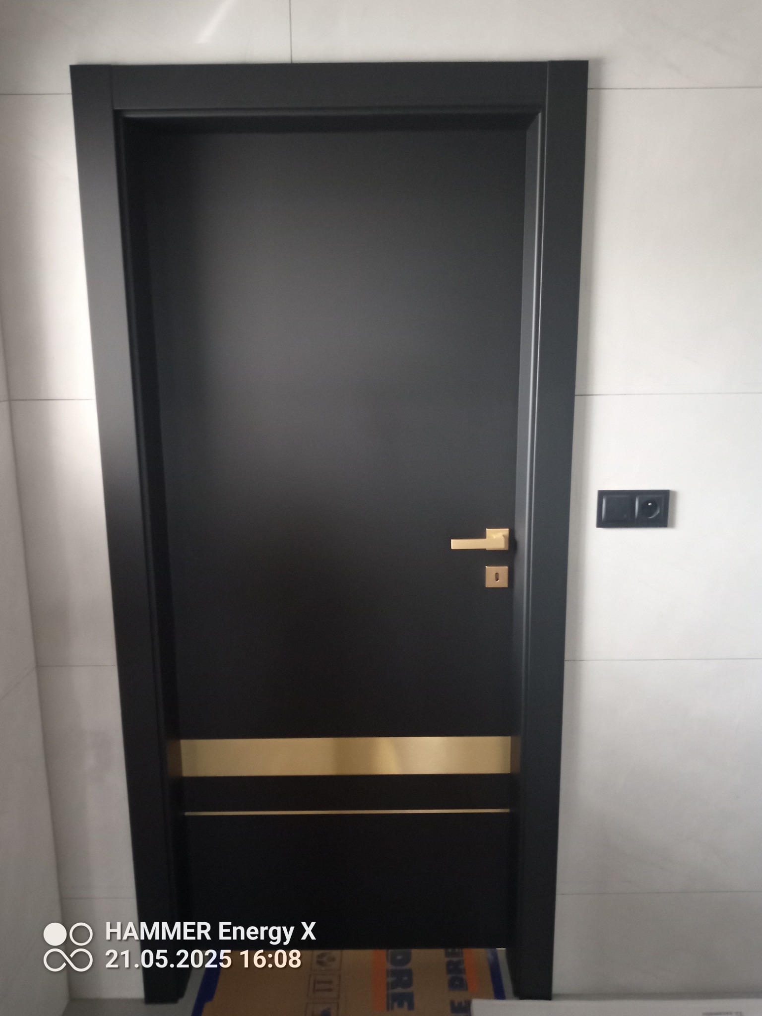 Czarne drzwi wewnętrzne z złotymi akcentami i klamką, zamontowane w białej ścianie z włącznikiem. Widoczny fragment kartonu na podłodze.