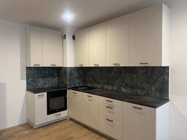 Nowoczesna kuchnia z białymi szafkami, czarnym blatem i marmurowym panelem ściennym. Minimalistyczne uchwyty, oświetlenie sufitowe. Ukończona realizacja.