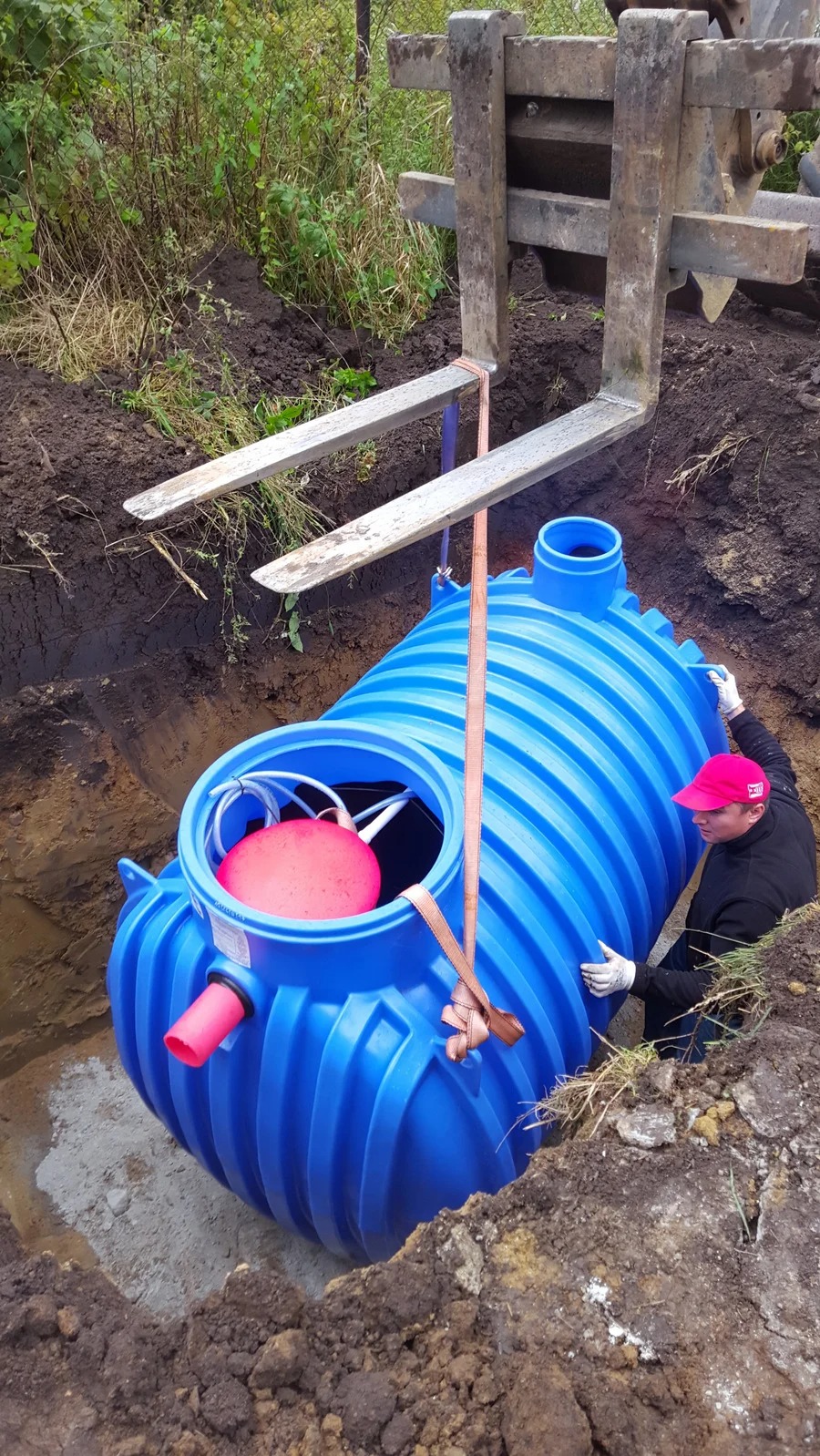 Montaż niebieskiego zbiornika z czerwonym pływakiem w wykopie, podtrzymywanego przez maszynę budowlaną. Pracownik w rękawicach i czapce asekuruje zbiornik.