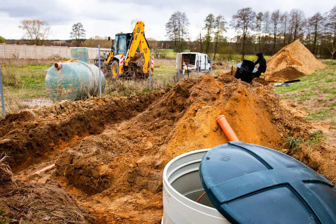Montaż przydomowej oczyszczalni ścieków: biały zbiornik z niebieską pokrywą na pierwszym planie, wykopany rów, koparka i pracownik w tle.