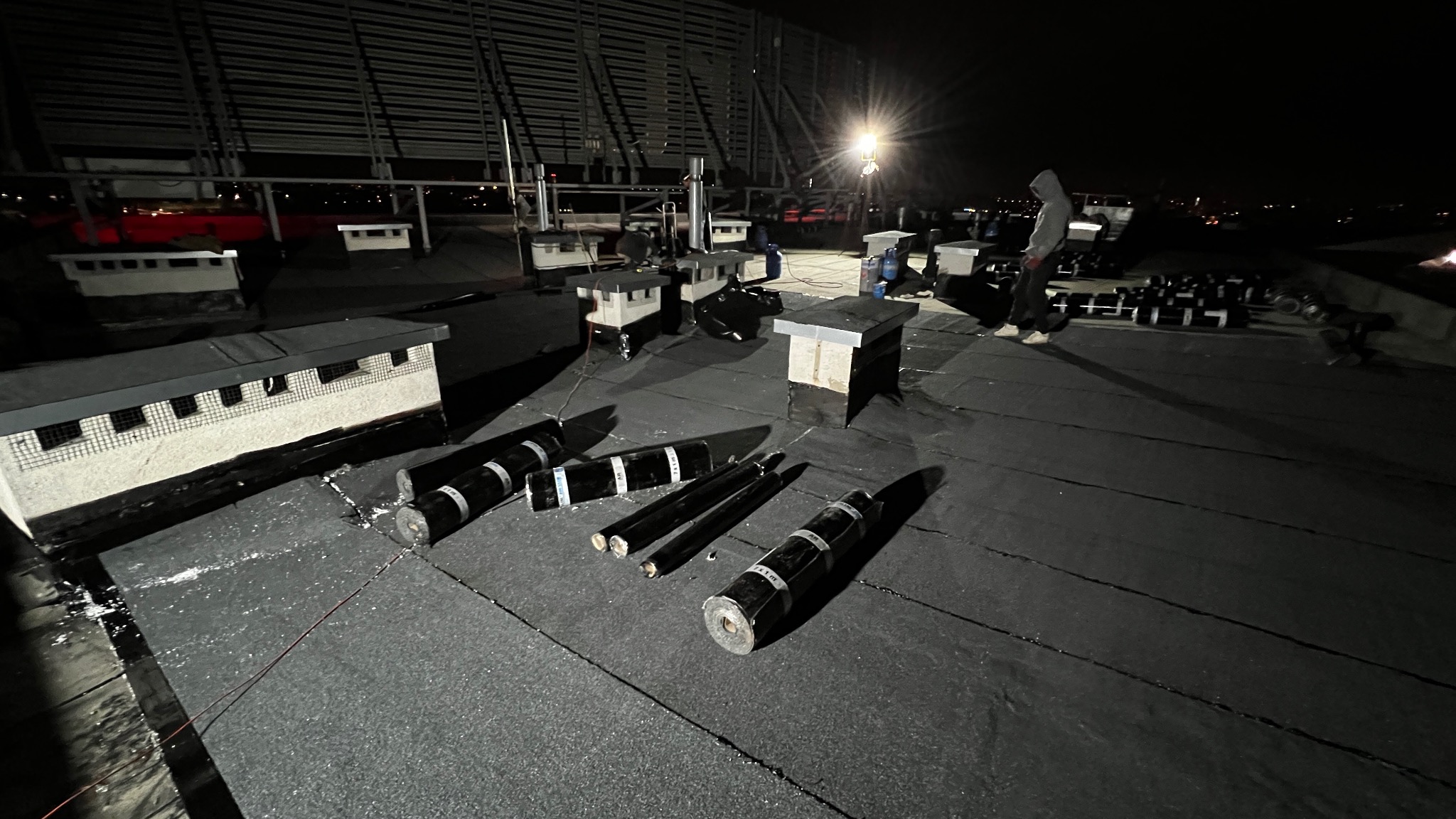 Nocne prace na dachu: rozłożone rolki papy bitumicznej, wentylatory i pracownik w kapturze w tle, oświetlone sztucznym światłem. Widok z góry.