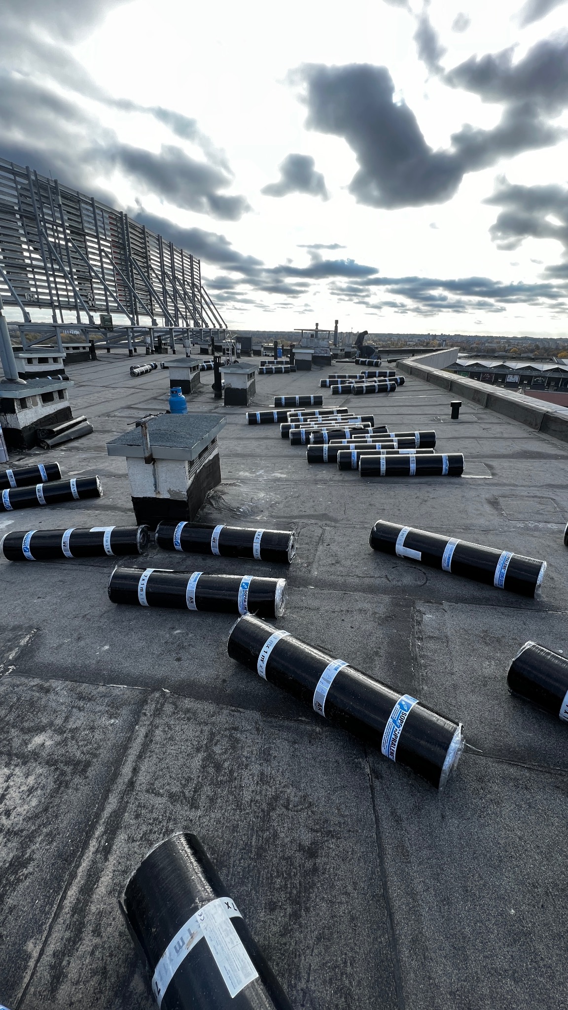 Rozłożone rolki papy termozgrzewalnej na płaskim dachu przed montażem, widoczne elementy wentylacji i konstrukcja wsporcza. Pochmurne niebo w tle.