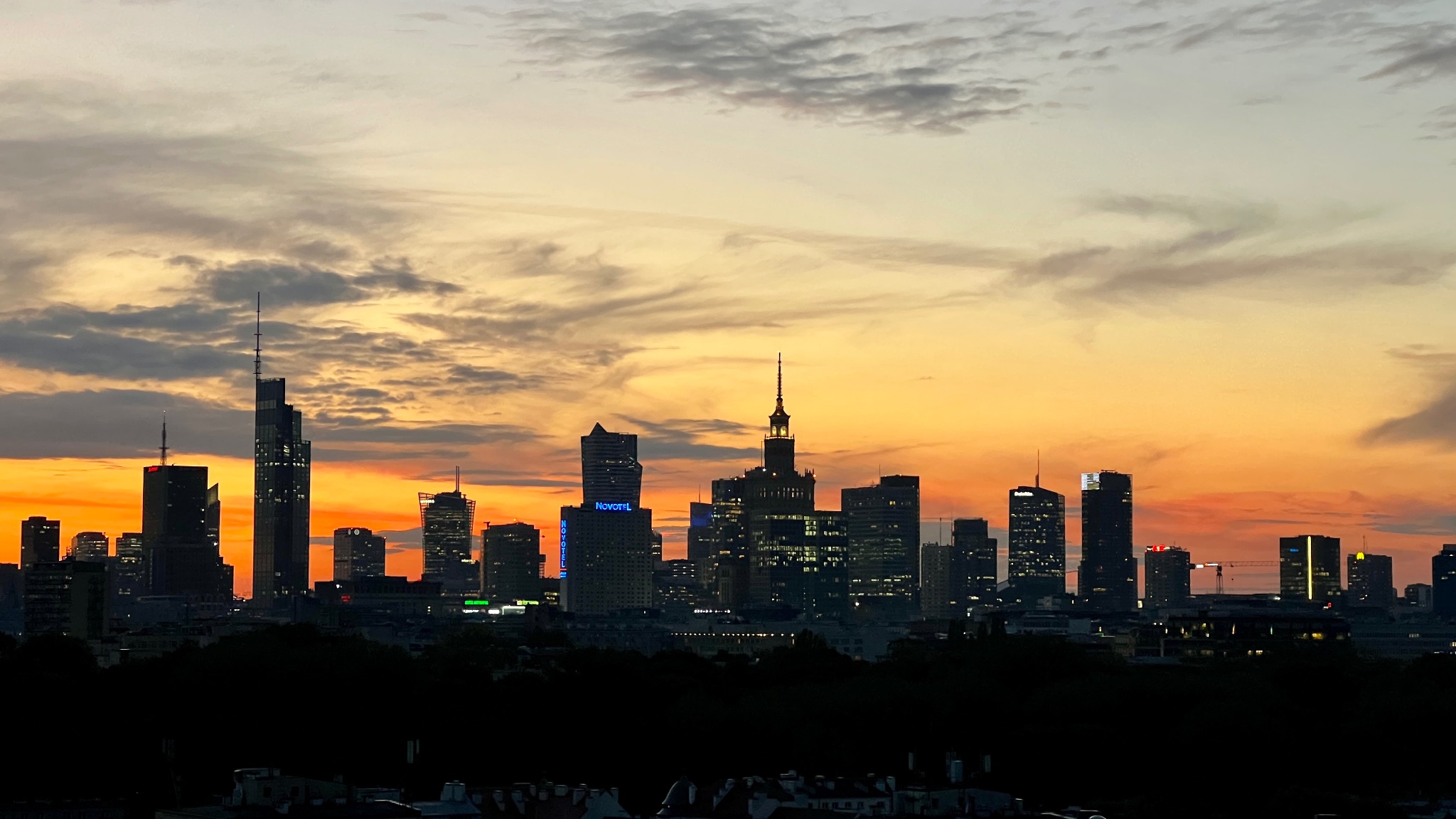 Panorama Warszawy o zmierzchu: sylwetki wieżowców na tle pomarańczowego nieba z chmurami. Widoczne oświetlone budynki i żuraw budowlany.