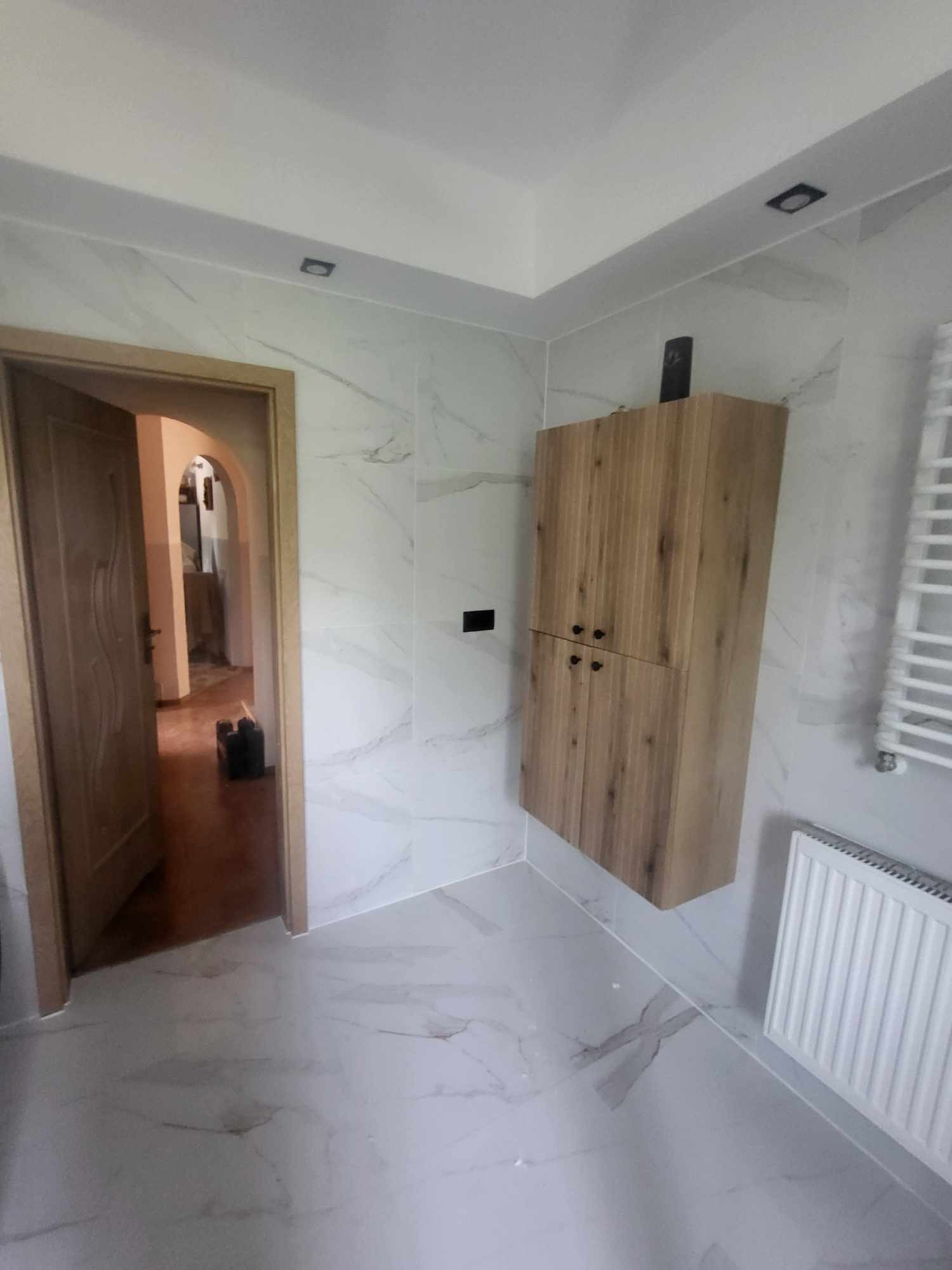 Wykończona łazienka z drewnianą szafką wiszącą, grzejnikiem i marmurowymi płytkami na ścianach i podłodze. Widok na otwarte drzwi do innego pomieszczenia.