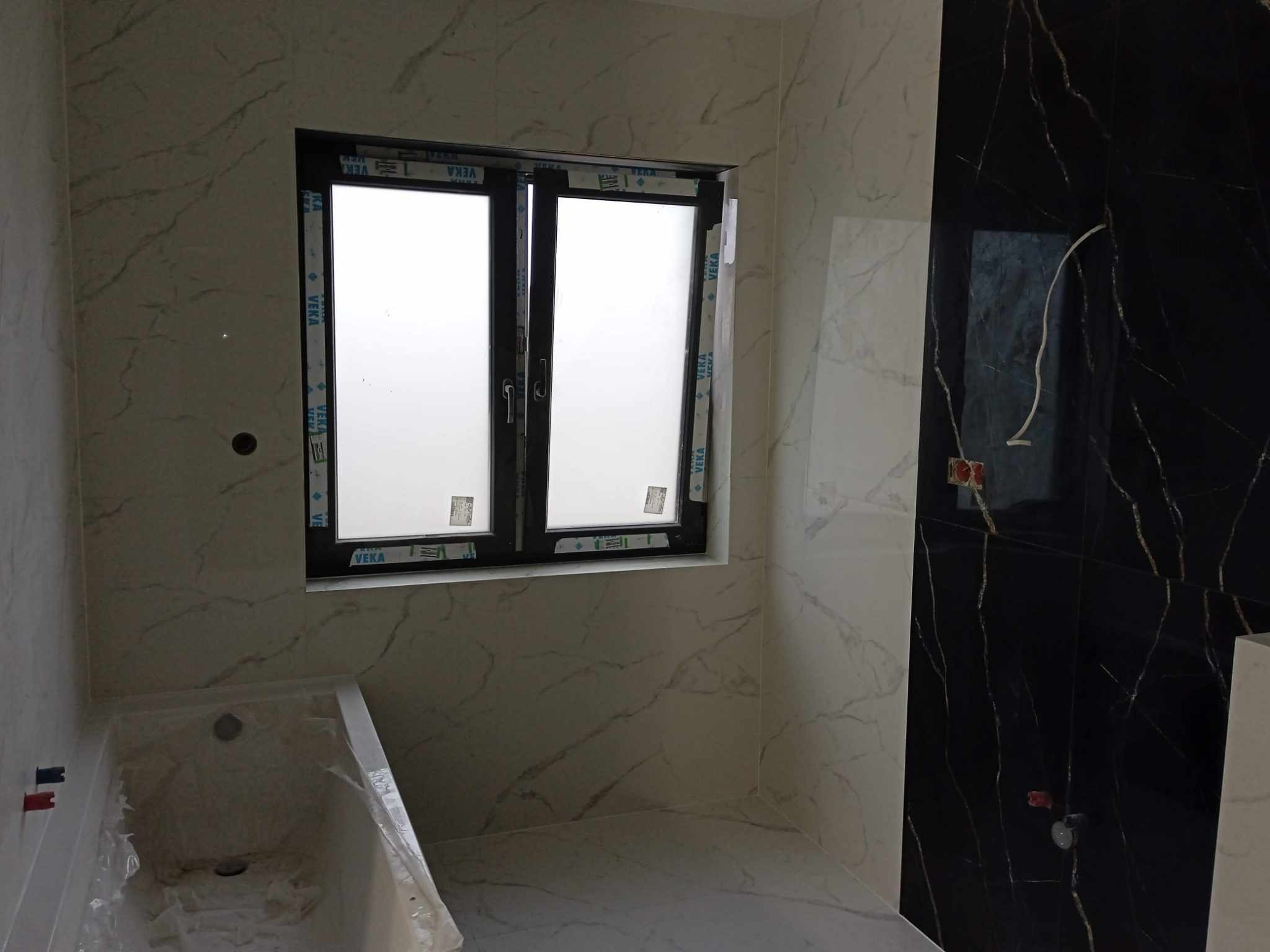 Wykończona łazienka z czarno-białymi płytkami imitującymi marmur, czarną ramą okienną i wanną zabezpieczoną folią. Widoczne przyłącza wodne.