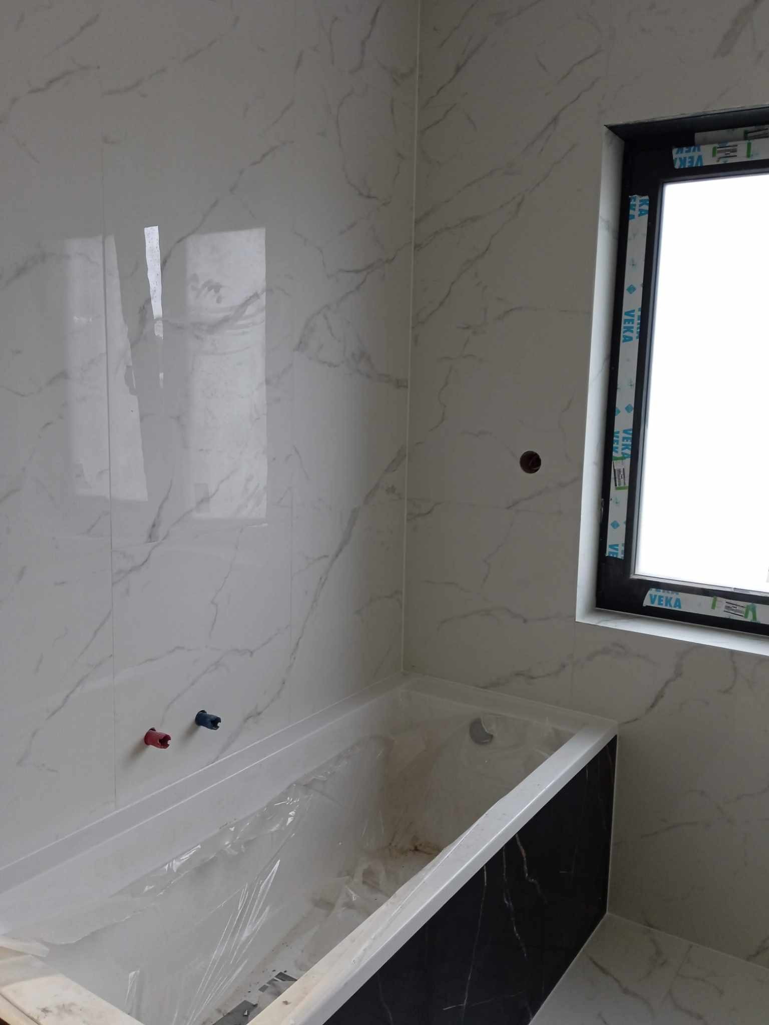 Biała wanna z czarnym panelem bocznym, zabezpieczona folią, w łazience z marmurowymi płytkami i oknem z czarną ramą. Widoczne wystające rury do baterii.