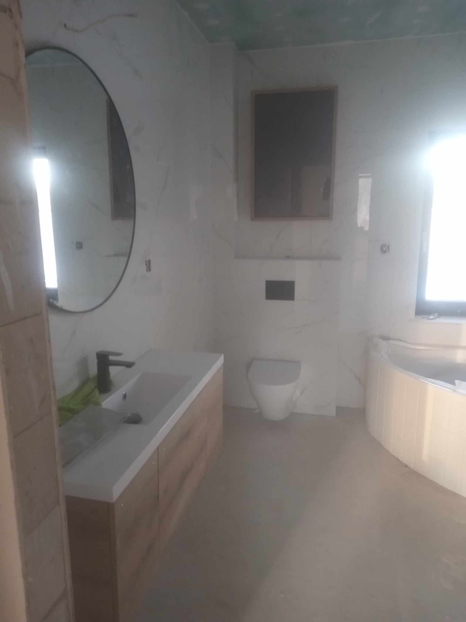 Wykończona łazienka z okrągłym lustrem, umywalką z szafką w kolorze drewna, toaletą i wanną. Białe marmurowe płytki na ścianach i podłodze. Minimalistyczny design.