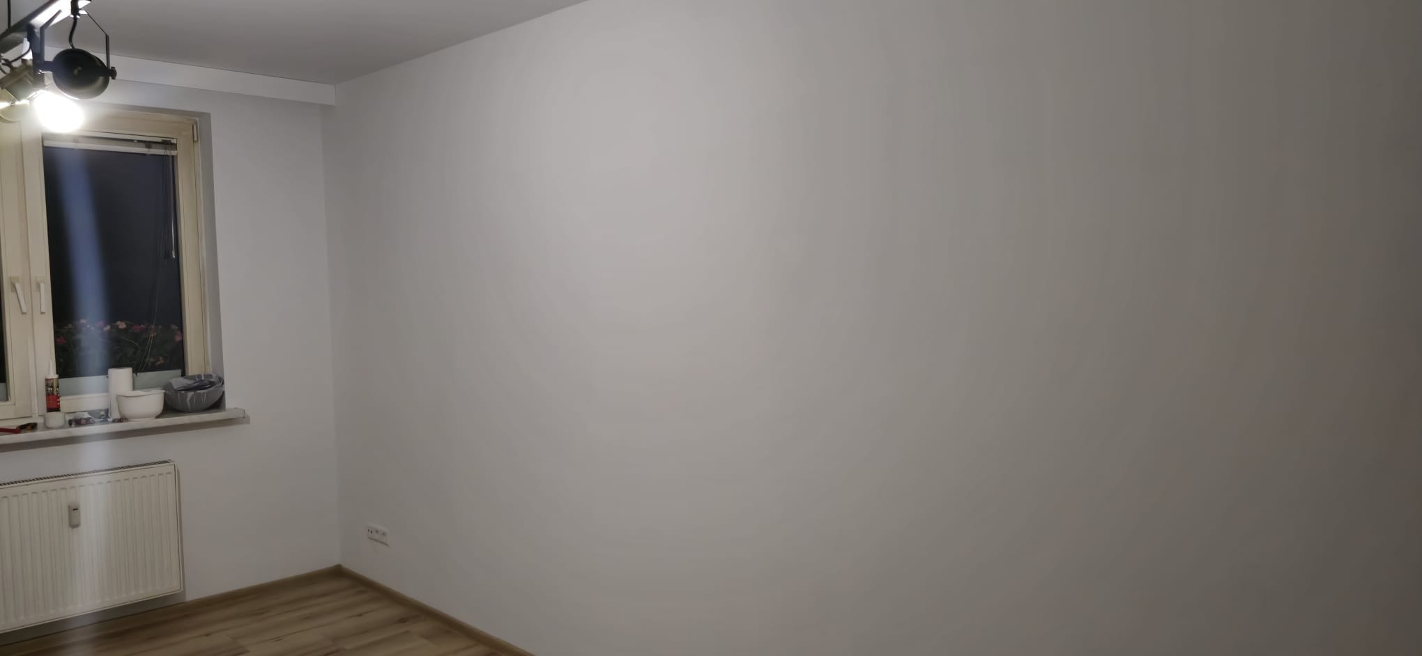 Jasne wnętrze po remoncie: gładkie, białe ściany, drewnopodobna podłoga, okno z widokiem na zieleń. Minimalistyczny design, czystość i porządek.