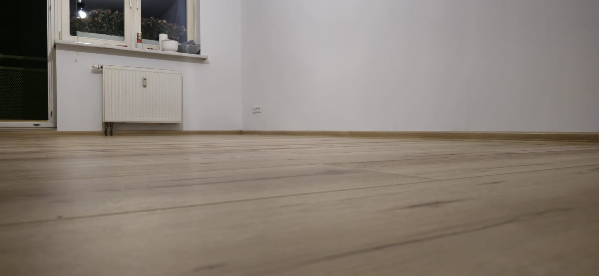 Jasna podłoga panelowa w pokoju po remoncie w Nowej Kuźni. Widoczny grzejnik, okno i białe ściany. Ujęcie z niskiej perspektywy podkreśla fakturę podłogi.