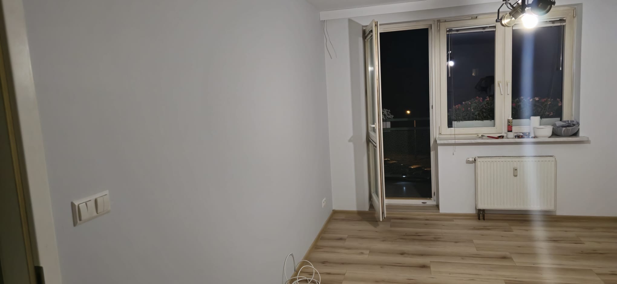 Jasne wnętrze pokoju po remoncie: świeżo pomalowane ściany w odcieniach szarości, drewniana podłoga, okno z widokiem na nocne miasto i biały grzejnik pod nim.