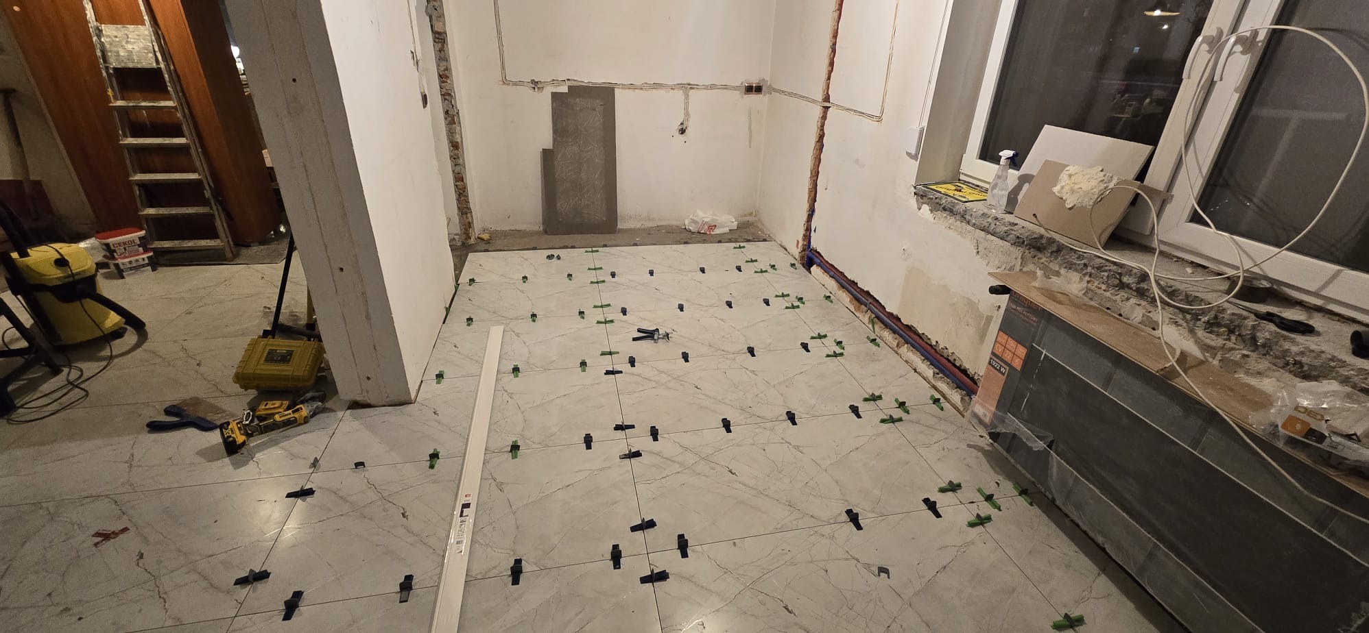 Układanie płytek podłogowych z użyciem klipsów poziomujących w trakcie prac. Widoczne narzędzia i materiały budowlane. Jasne płytki imitujące marmur.