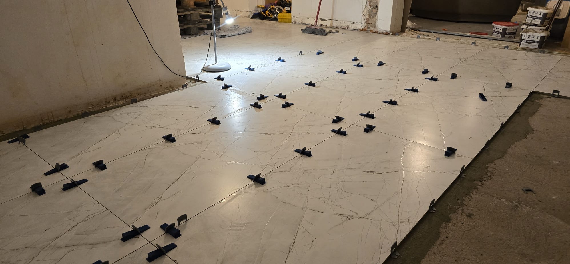 Układanie płytek podłogowych z użyciem systemu poziomowania. Widoczne krzyżyki dystansowe i kliny. W tle lampa robocza i materiały budowlane.