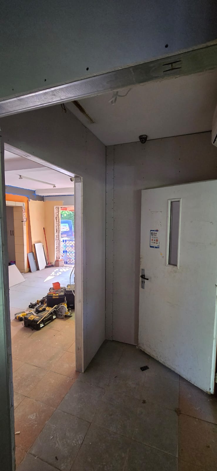 Wnętrze w trakcie prac: ściany obłożone płytami gipsowo-kartonowymi, widoczne narzędzia i materiały budowlane. Drzwi z małym okienkiem i kamera przemysłowa na suficie.