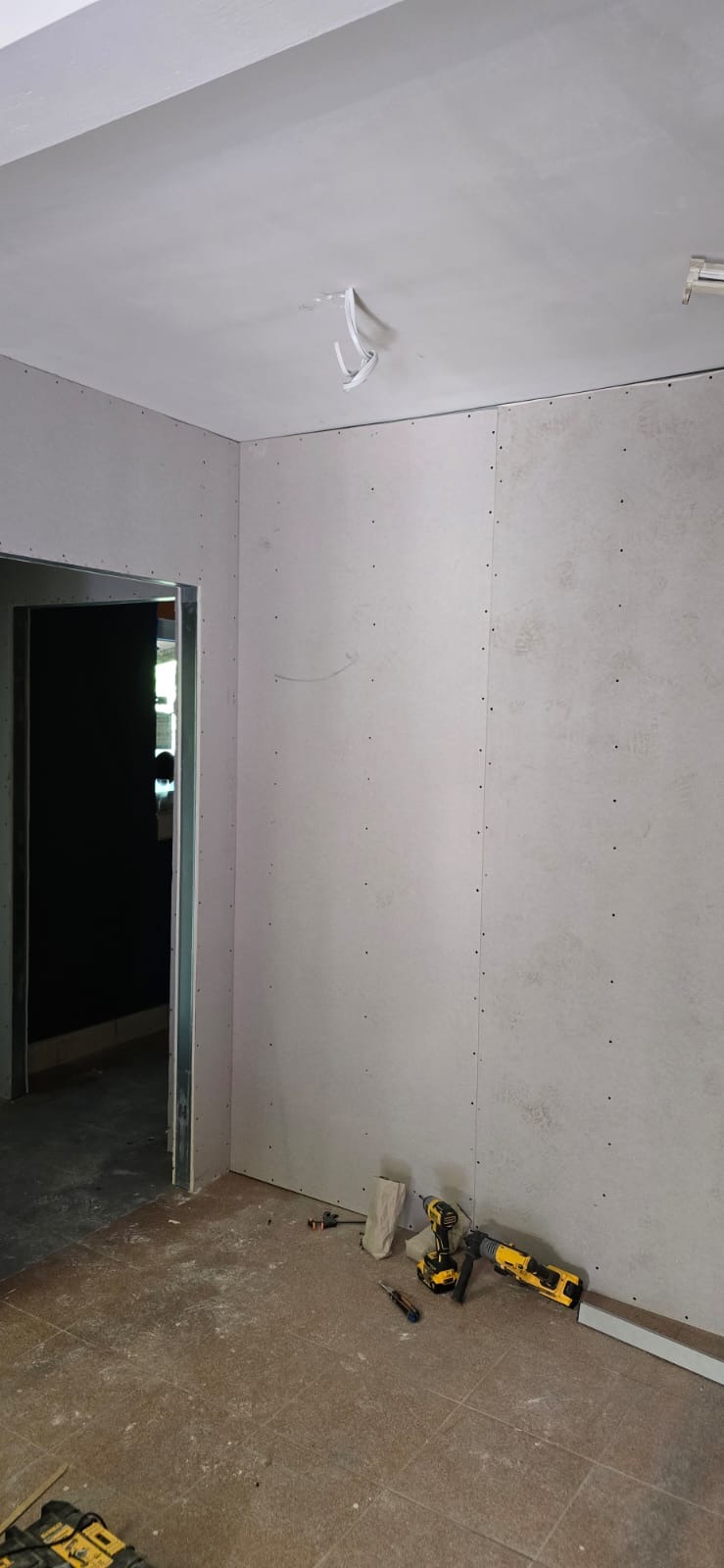 Wnętrze w trakcie montażu płyt karton-gips na ścianach i suficie. Widoczne narzędzia pracy: wkrętarka, poziomica. Otwór drzwiowy z metalową ościeżnicą.