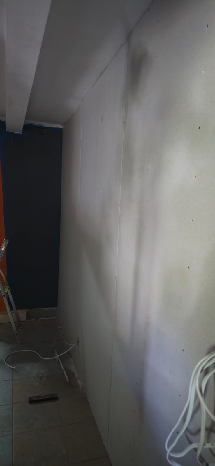 Ściana z płyt kartonowo-gipsowych w trakcie montażu, widoczne łączenia płyt, fragment drabiny i przewody na podłodze. Ujęcie z dołu, perspektywa zbieżna.