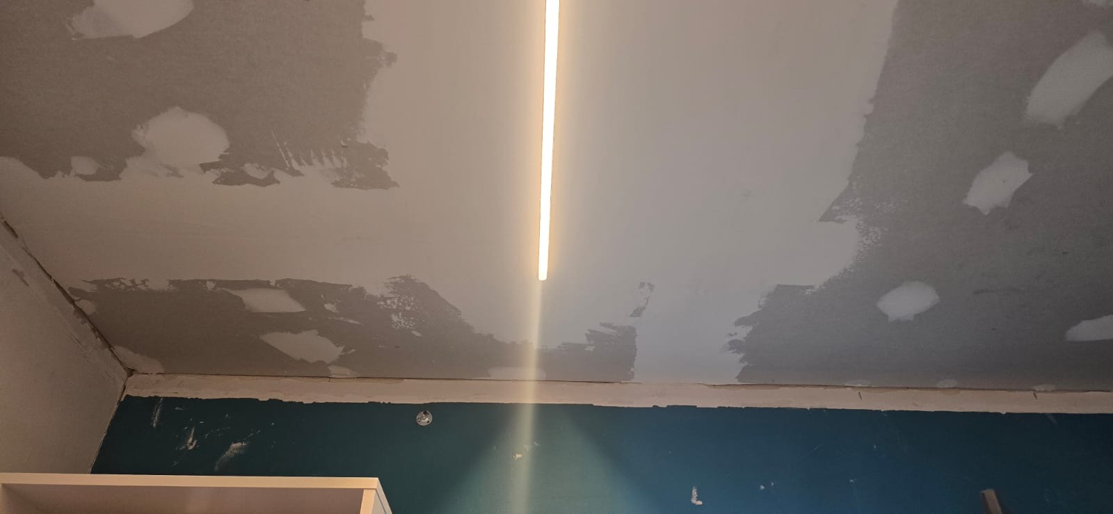 Nierówna powierzchnia sufitu po szpachlowaniu, z widoczną linią oświetlenia LED. Fragment niebieskiej ściany i białego mebla w dolnej części kadru. Etap prac wykończeniowych.