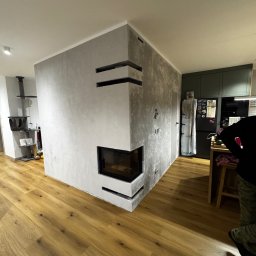 Rafał Dzionek - Nowoczesny salon z kominkiem obłożonym szarym tynkiem dekoracyjnym, drewniana podłoga, widok na kuchnię z zielonymi szafkami. Minimalistyczny design.