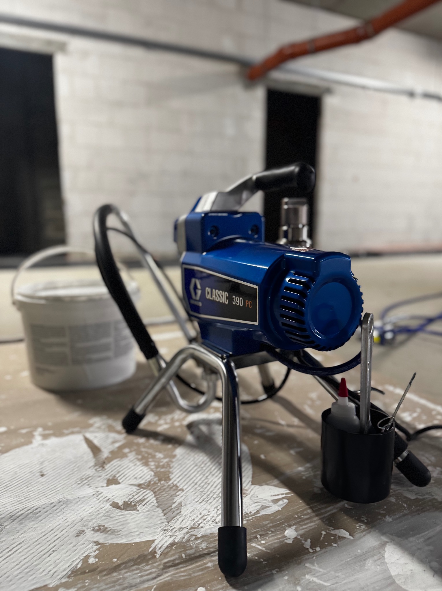 Niebieski agregat malarski Graco Classic 390 PC na podłodze z resztkami farby, obok wiadro z farbą. W tle surowa ściana i instalacje. Sprzęt do malowania.