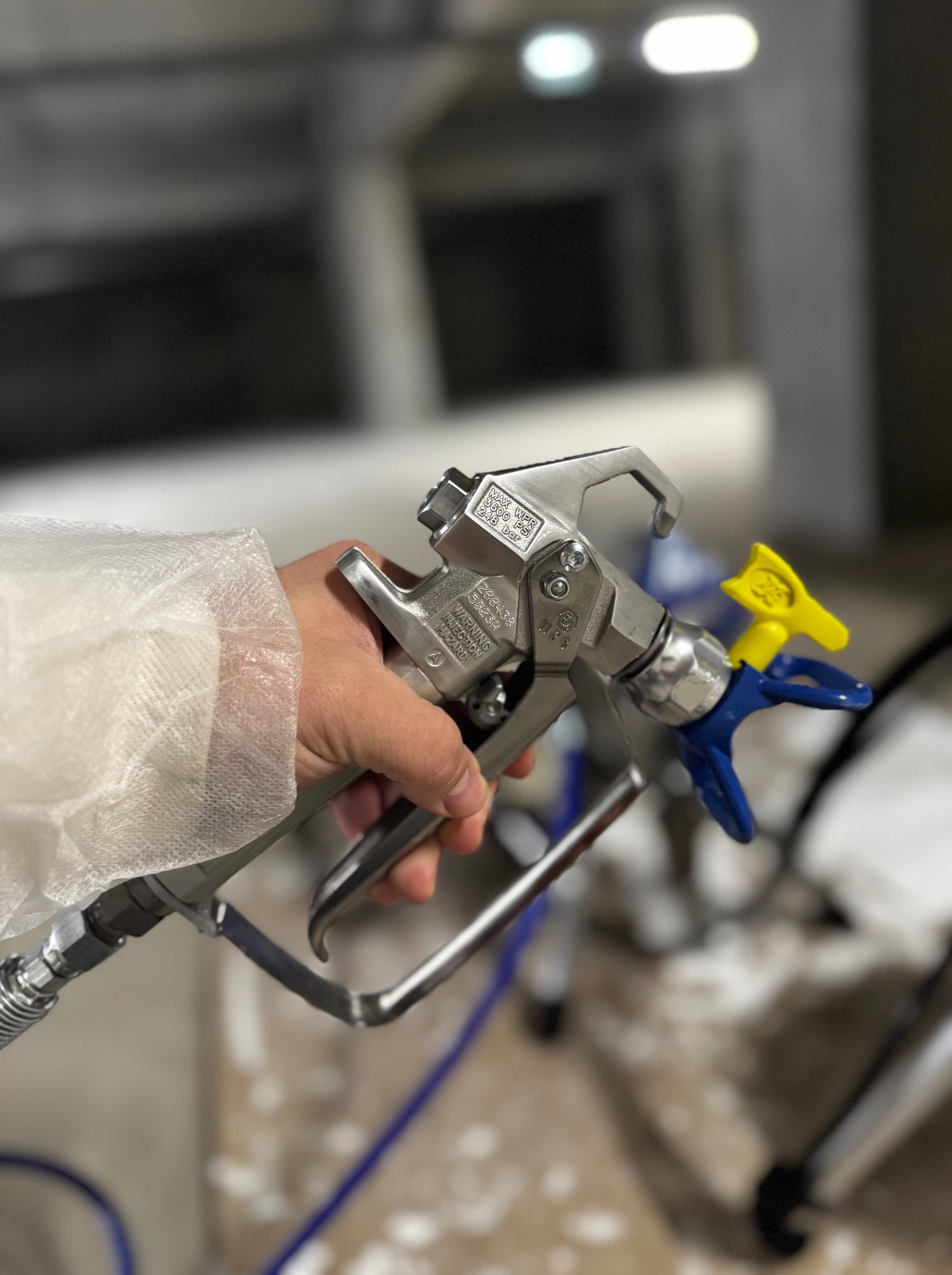 Dłoń w kombinezonie ochronnym trzyma pistolet natryskowy do malowania, widoczne detale dyszy i węża, tło rozmyte.