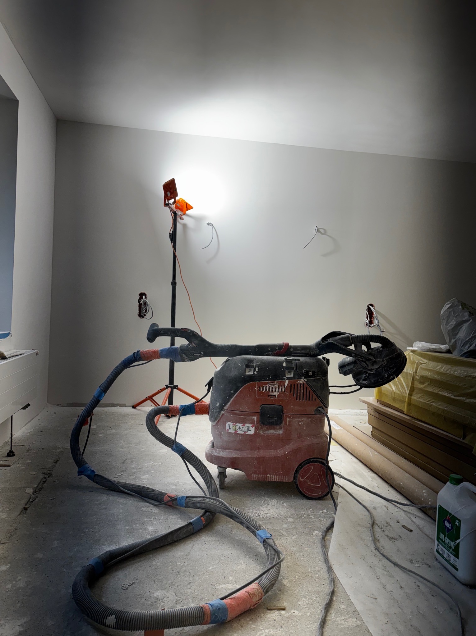 Pokój w trakcie szpachlowania: szlifierka do gładzi na odkurzaczu przemysłowym, lampa oświetleniowa, ściany przygotowane do malowania, widoczne puszki elektryczne.