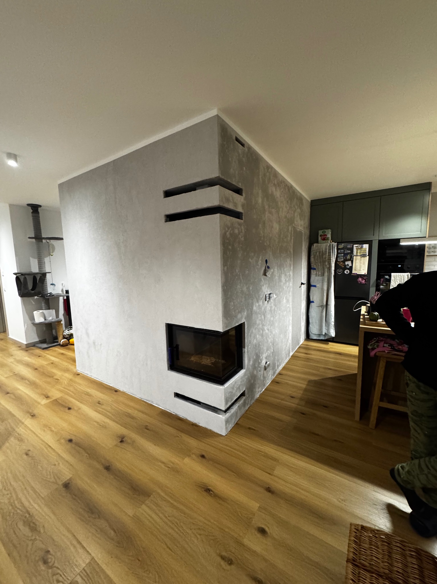Nowoczesny salon z kominkiem obłożonym szarym tynkiem dekoracyjnym, drewniana podłoga, widok na kuchnię z zielonymi szafkami. Minimalistyczny design.